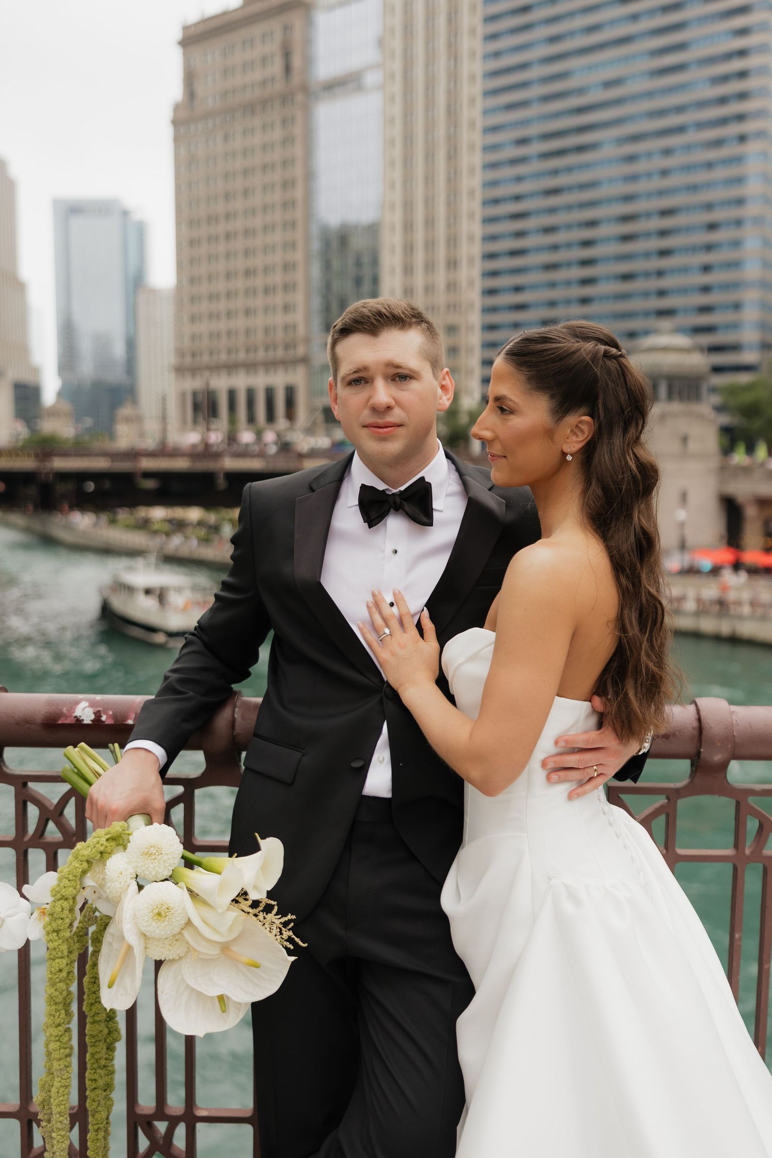 Bride and groom pose on a Chicago Riverwalk bridge, she in a strapless gown, he in a black tuxedo holding white florals.