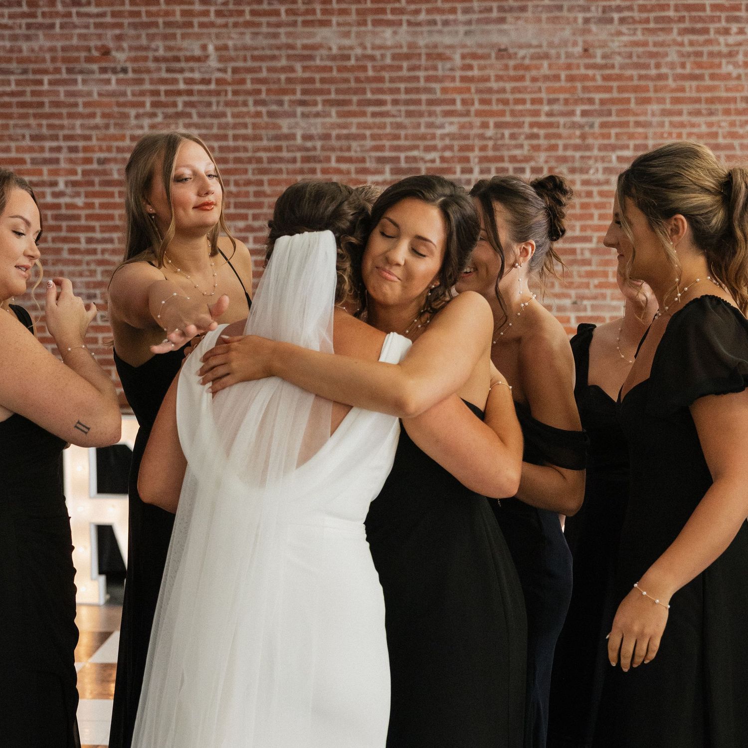 Wedding party wearing black dresses and white gowns share joyful moments against a brick wall backdrop.