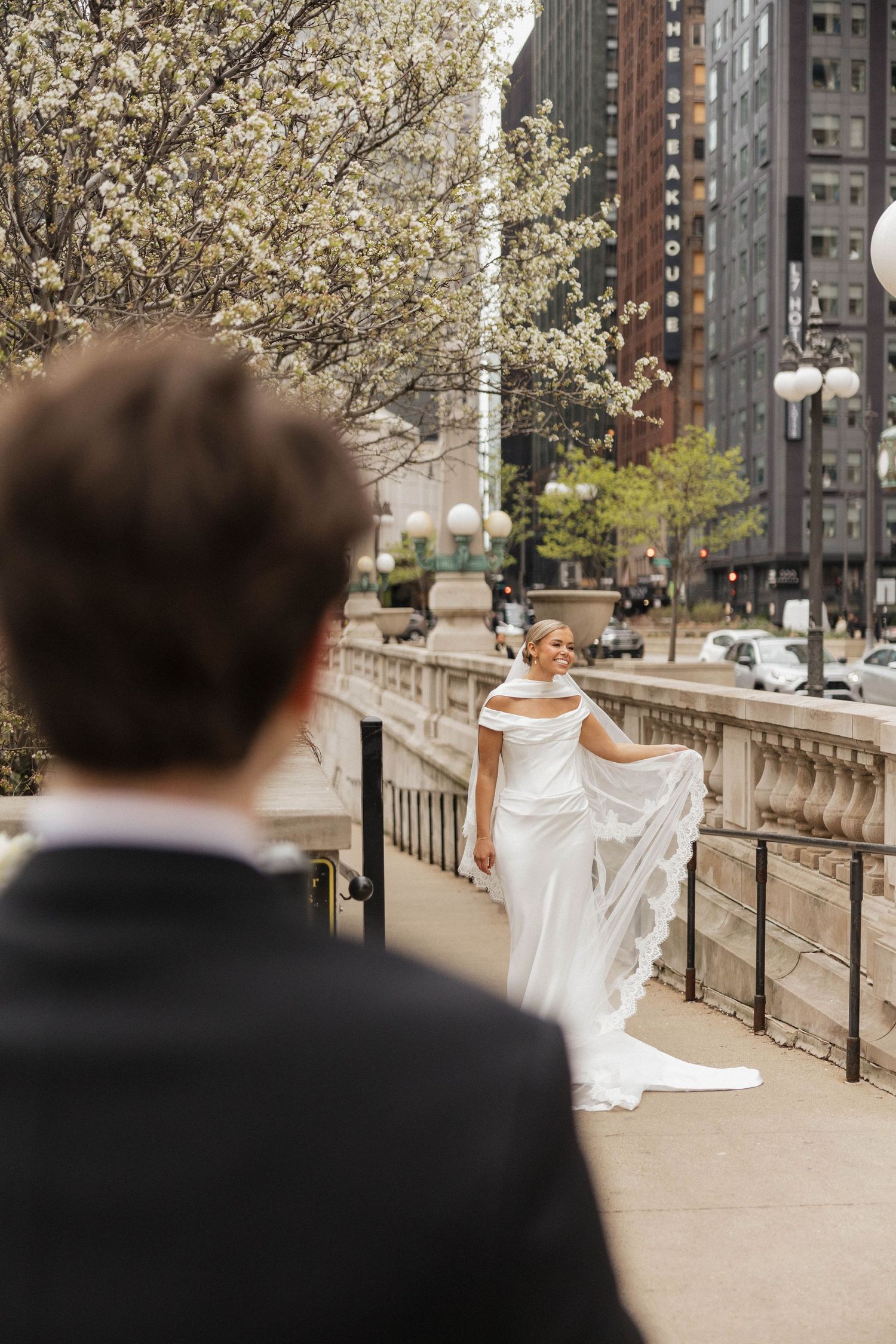 Bride in white off-shoulder gown poses on urban bridge while groom watches, with blooming trees and city skyline backdrop.