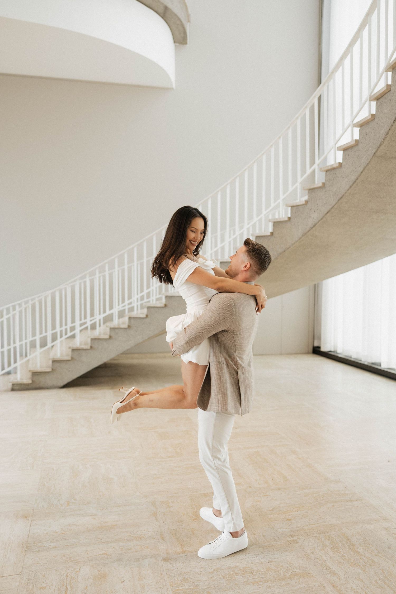 A man lifts a woman in a bright, elegant space with a curved staircase, both dressed in white and beige.
