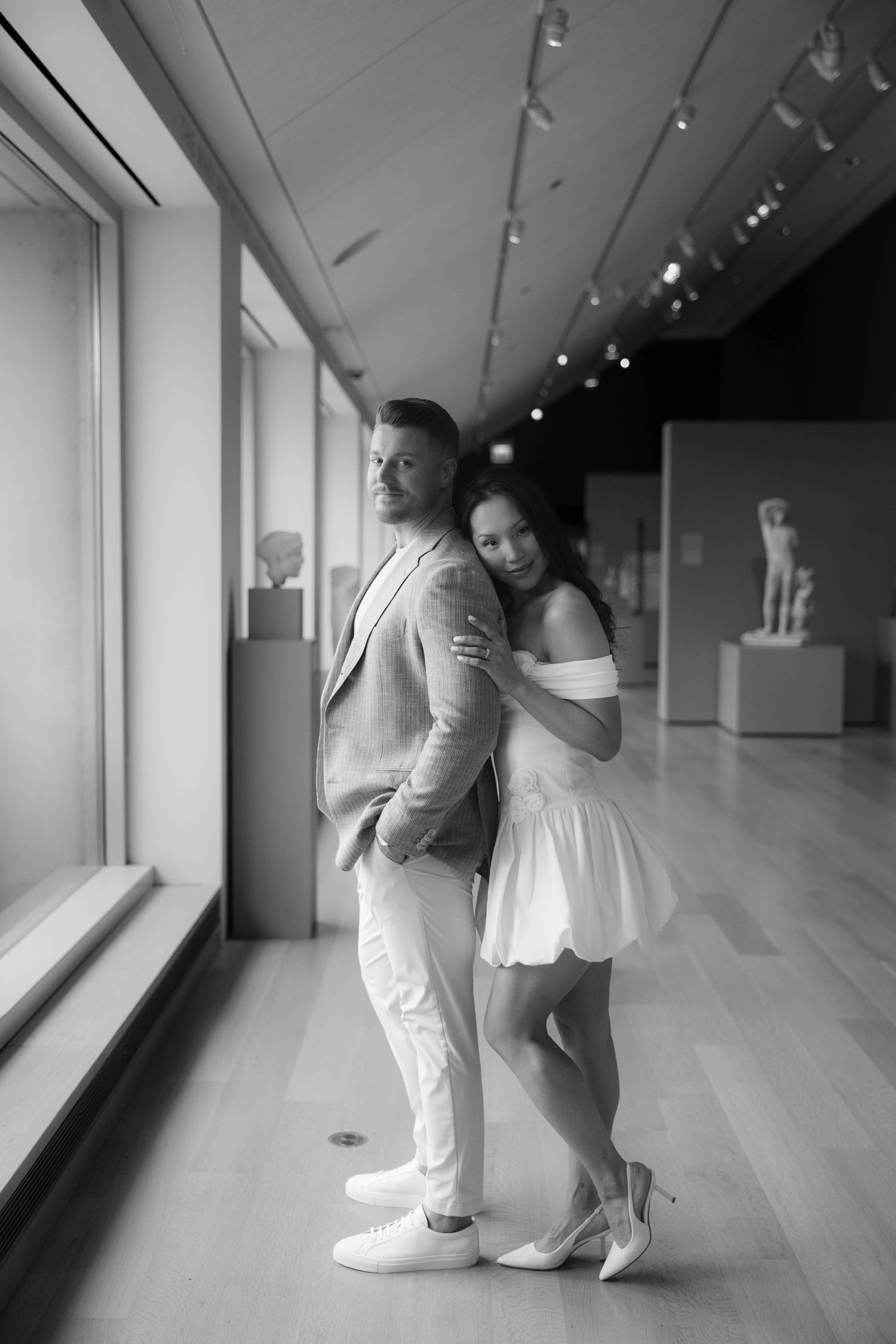 Black and white photo of a couple posing in a museum gallery, woman in white dress leaning on mans back.