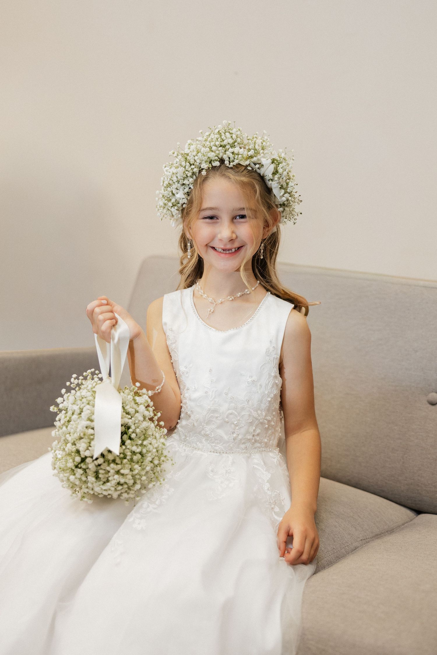 Smiling flower girl in white lace dress and baby's breath floral crown holds a round bouquet while seated on a gray sofa.