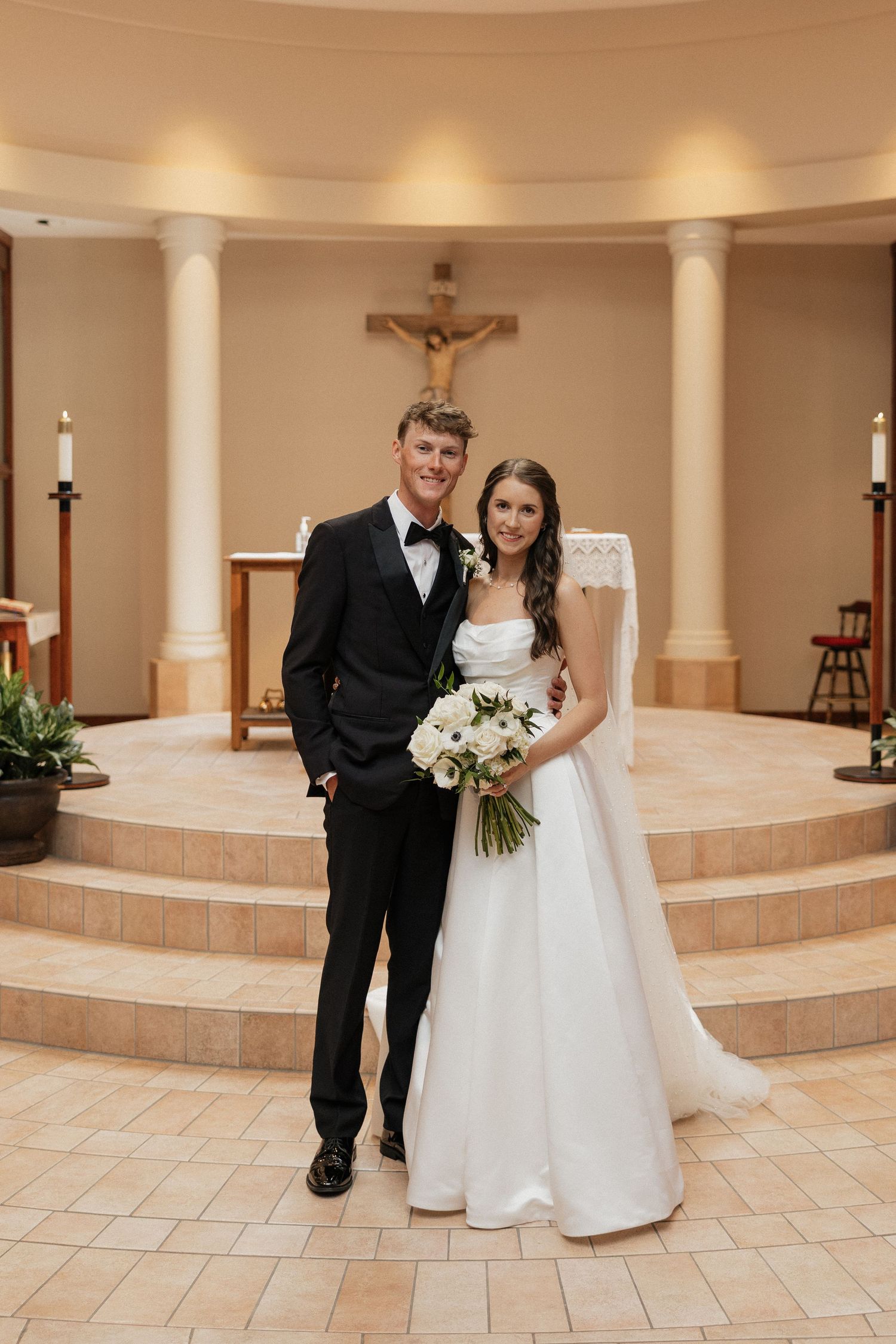 Bride and groom pose together at church altar, she holds white bouquet in strapless gown, he wears black tuxedo.
