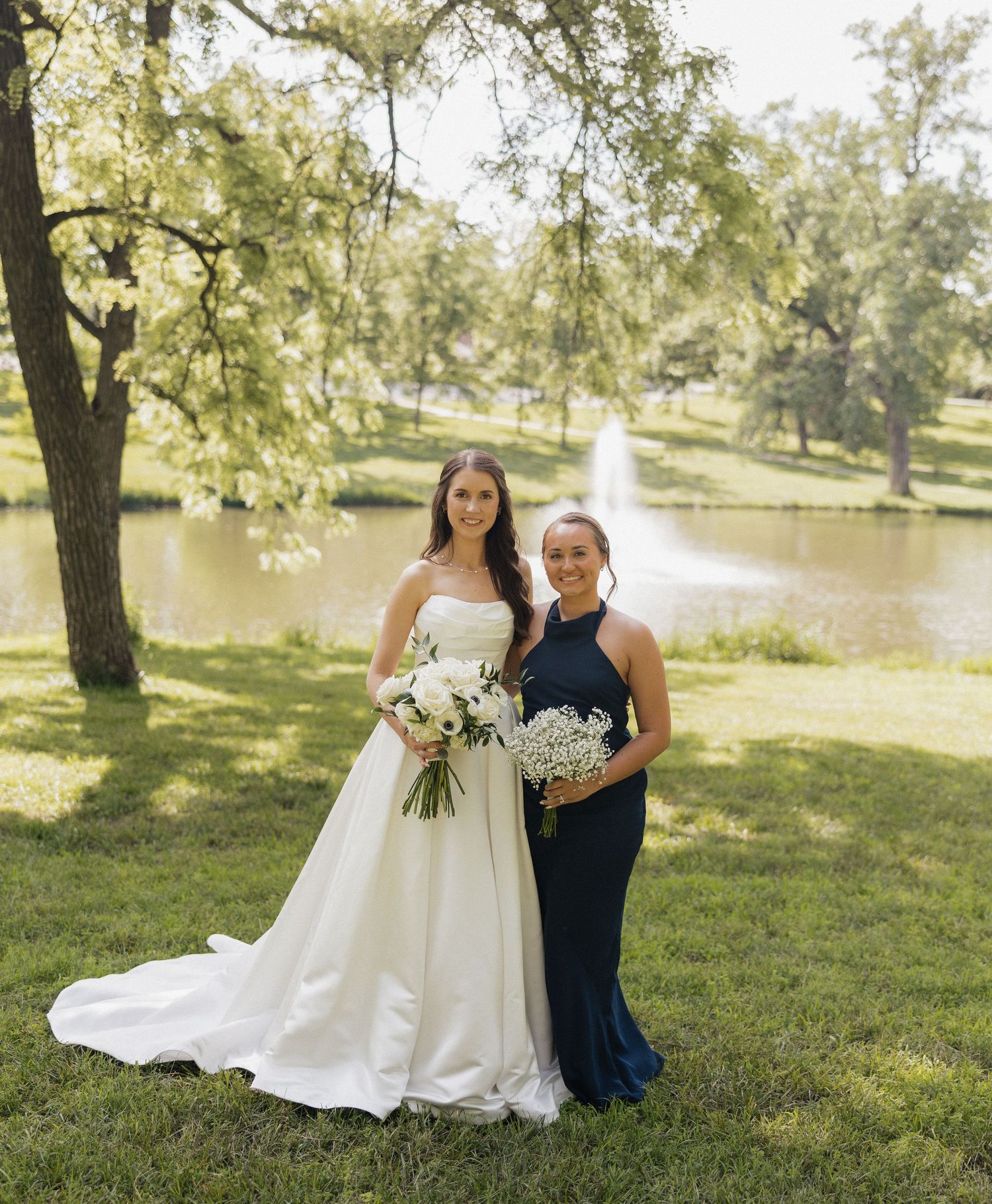 Bride in white gown and bridesmaid in navy dress holding bouquets outdoors near a pond with fountain.