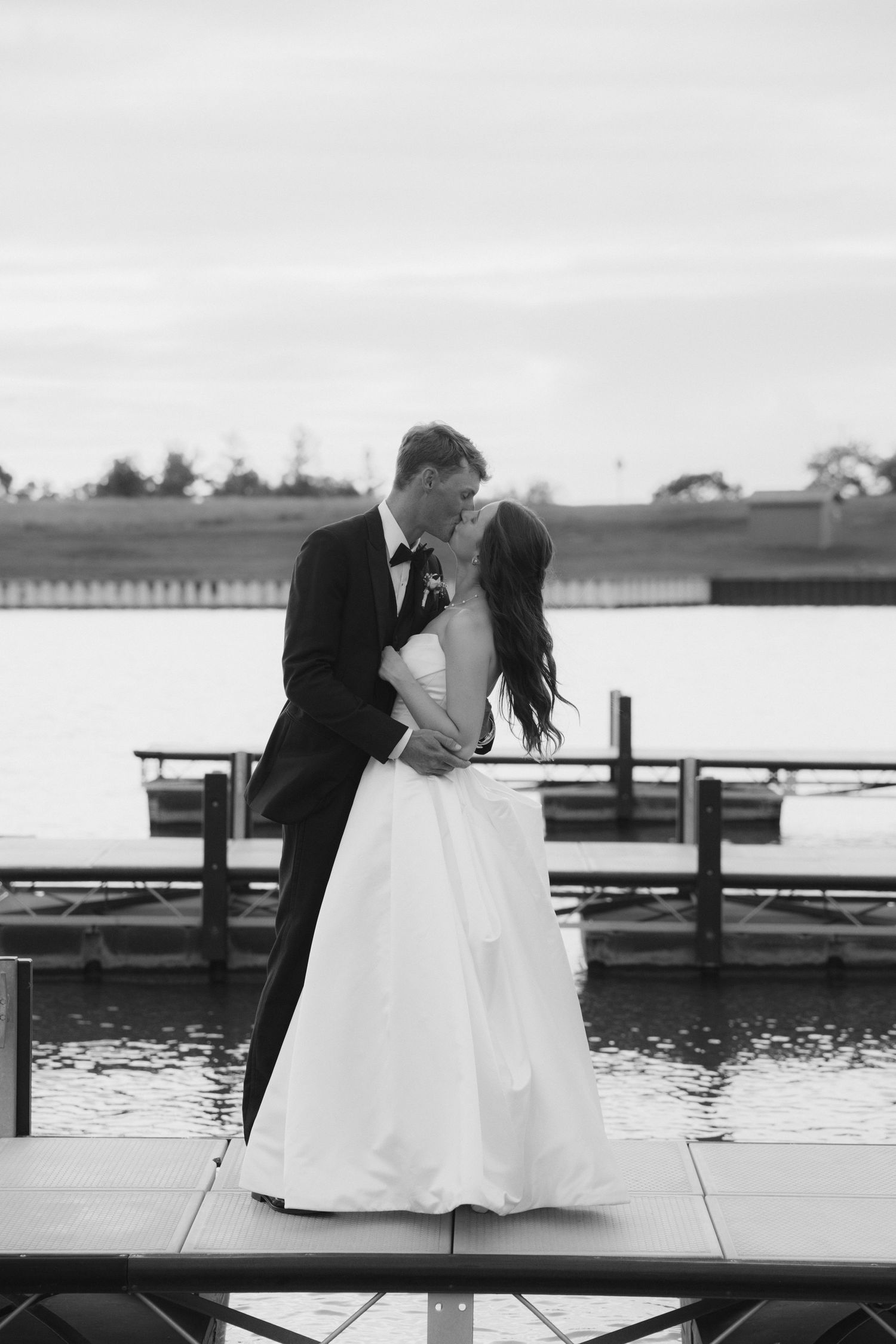 Black and white photo of a bride and groom sharing a kiss on a dock by the water in wedding attire.