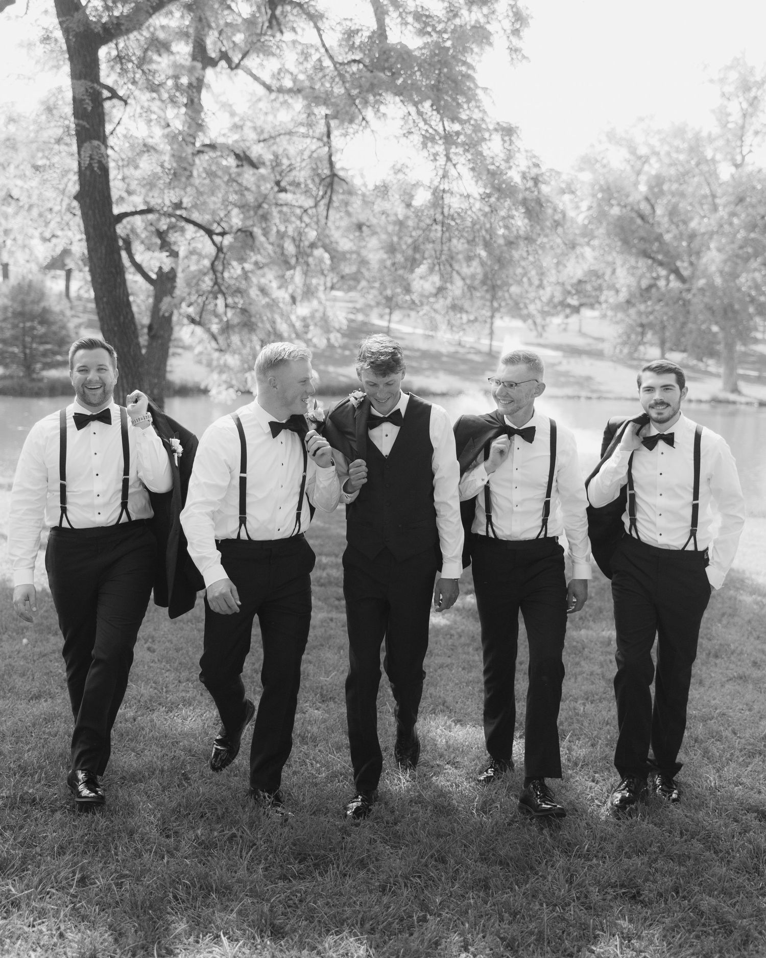 Five groomsmen in black tuxedos with bow ties and suspenders walk together outdoors in a classic black and white wedding photo.