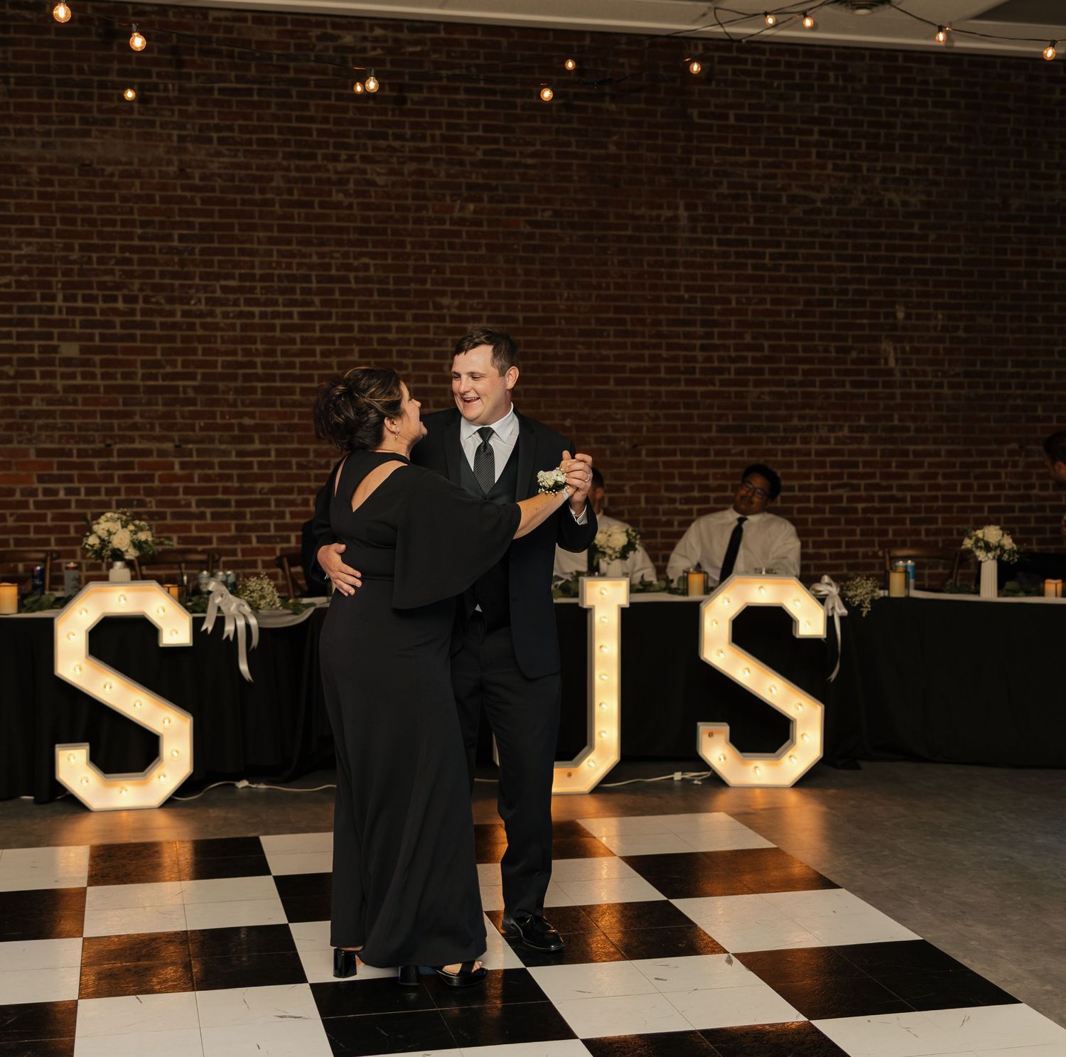 Couples dancing on black and white checkered floor with illuminated letters in background.