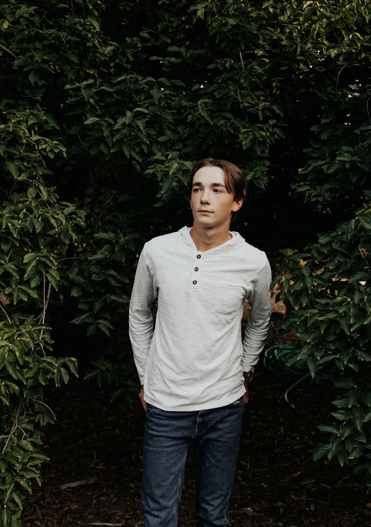 Portrait series against dark foliage wearing white henley shirt and jeans.