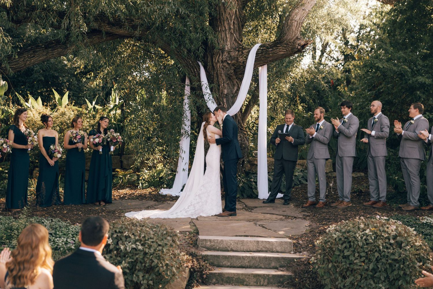 An outdoor wedding ceremony with guests seated under trees.