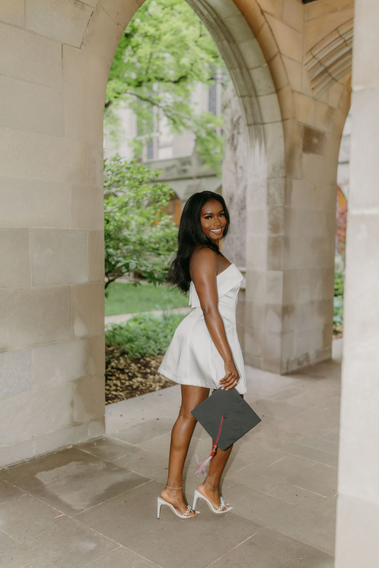 Graduate in white dress and heels poses under stone archway during outdoor photo shoot.