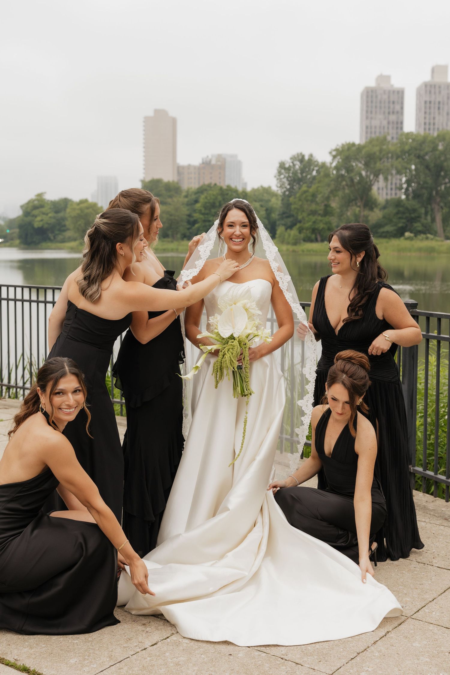 Bride in white gown holds bouquet as bridesmaids in black dresses adjust her veil and train outdoors near a city park.