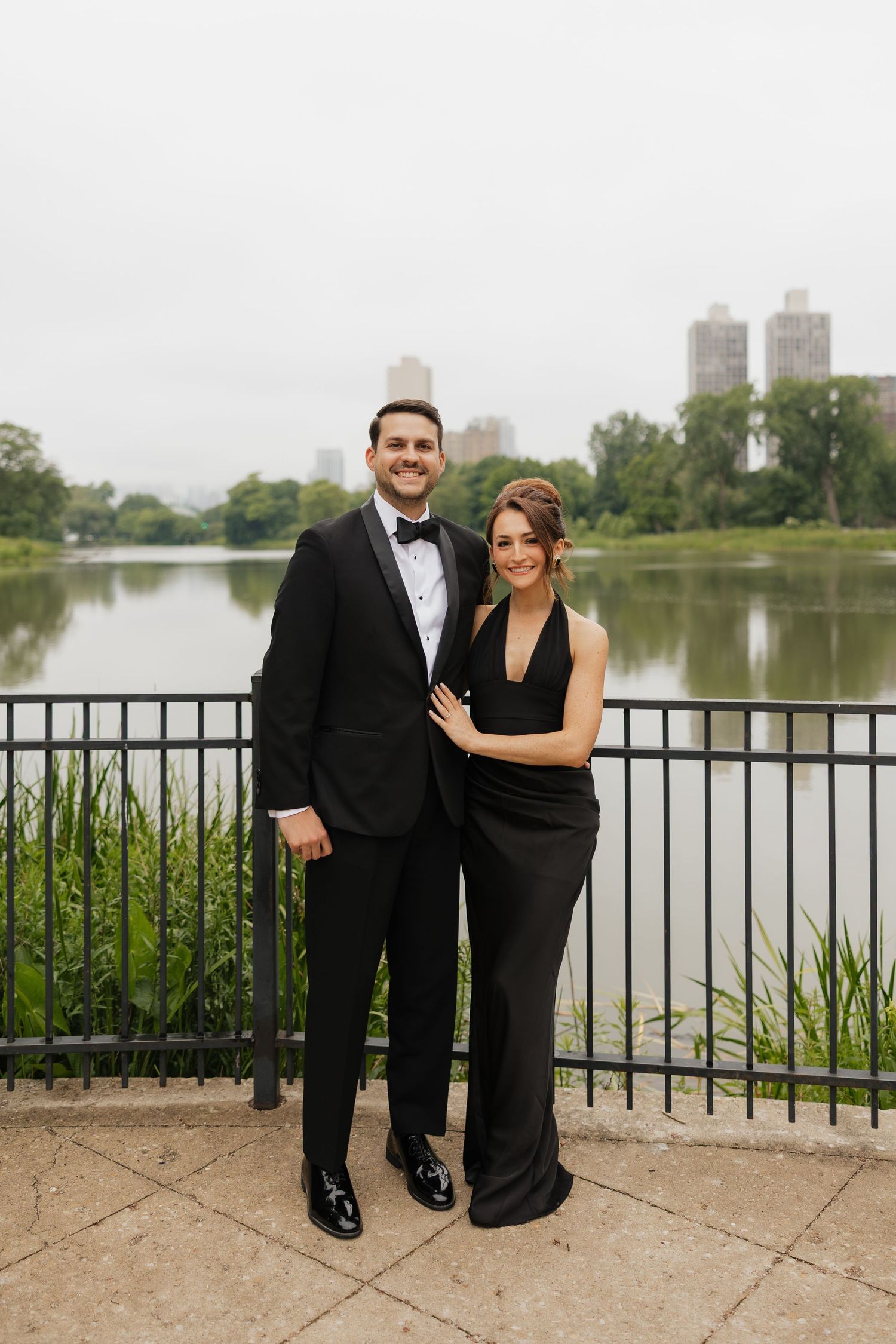 Couple in matching black formal attire posing by a railing with a city lake backdrop.