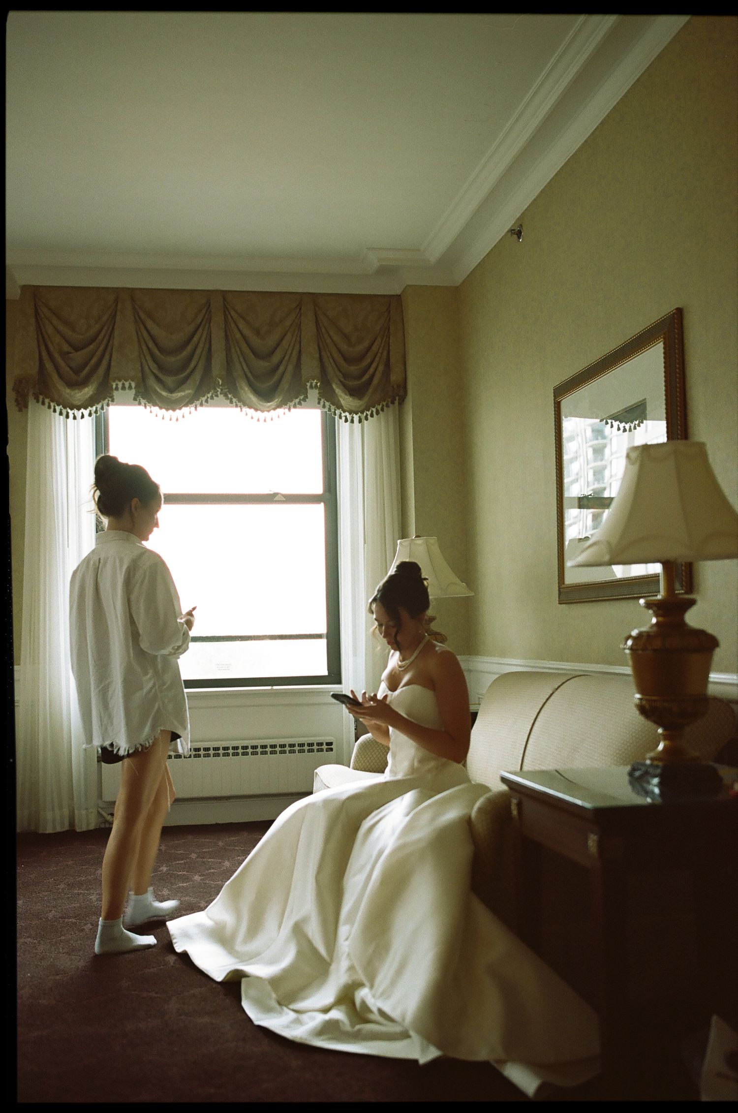 Bride in white gown sits in hotel room while bridesmaid checks phone near window on wedding day.