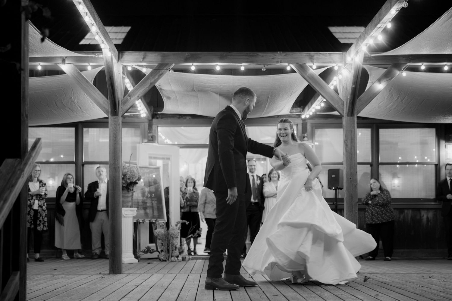 Bride in a flowing white gown and groom in a dark suit share a first dance on a wooden floor as wedding guests watch under string lights.