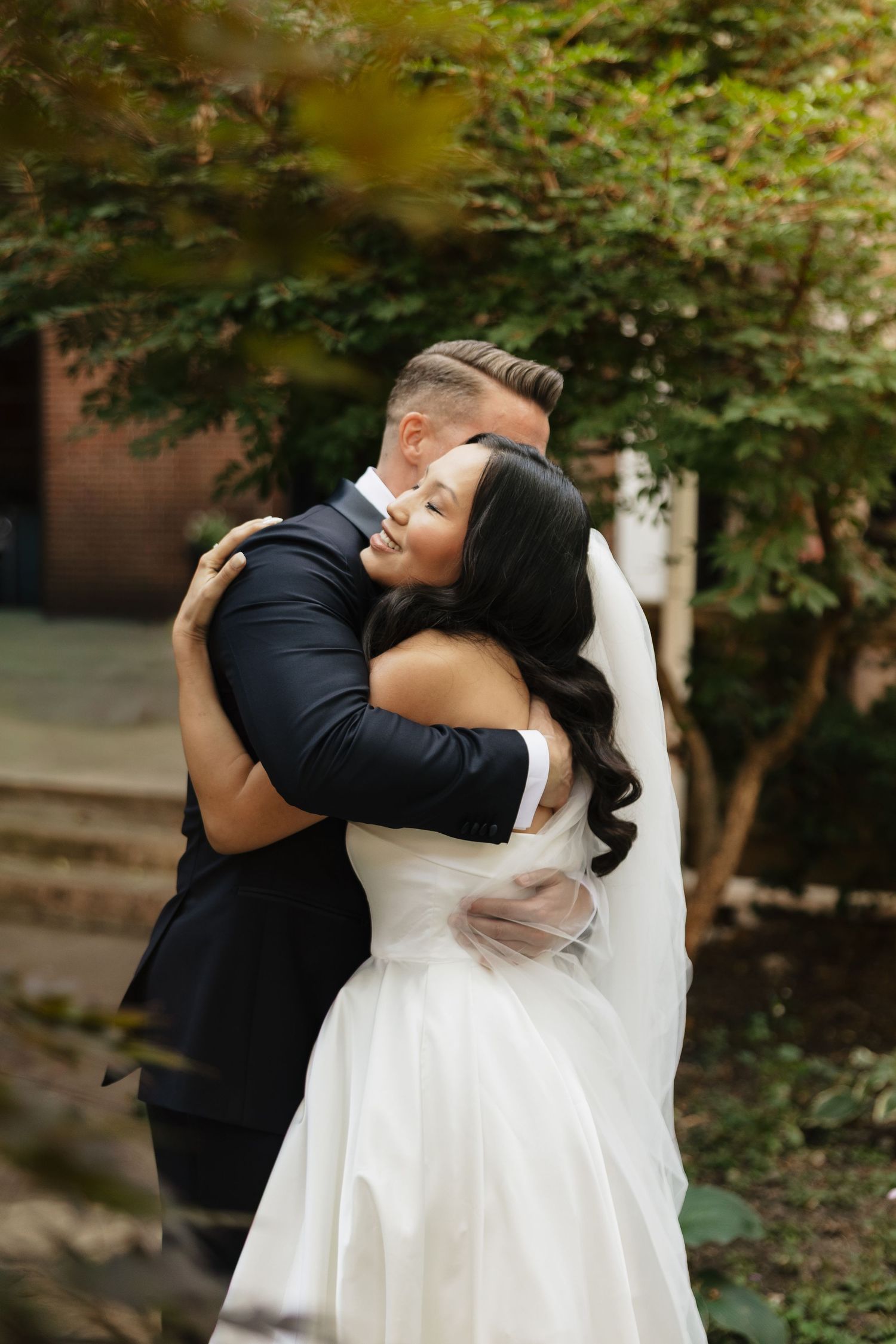 A bride in a white strapless gown and veil shares a warm, joyful embrace with a groom in a dark suit beneath green trees.