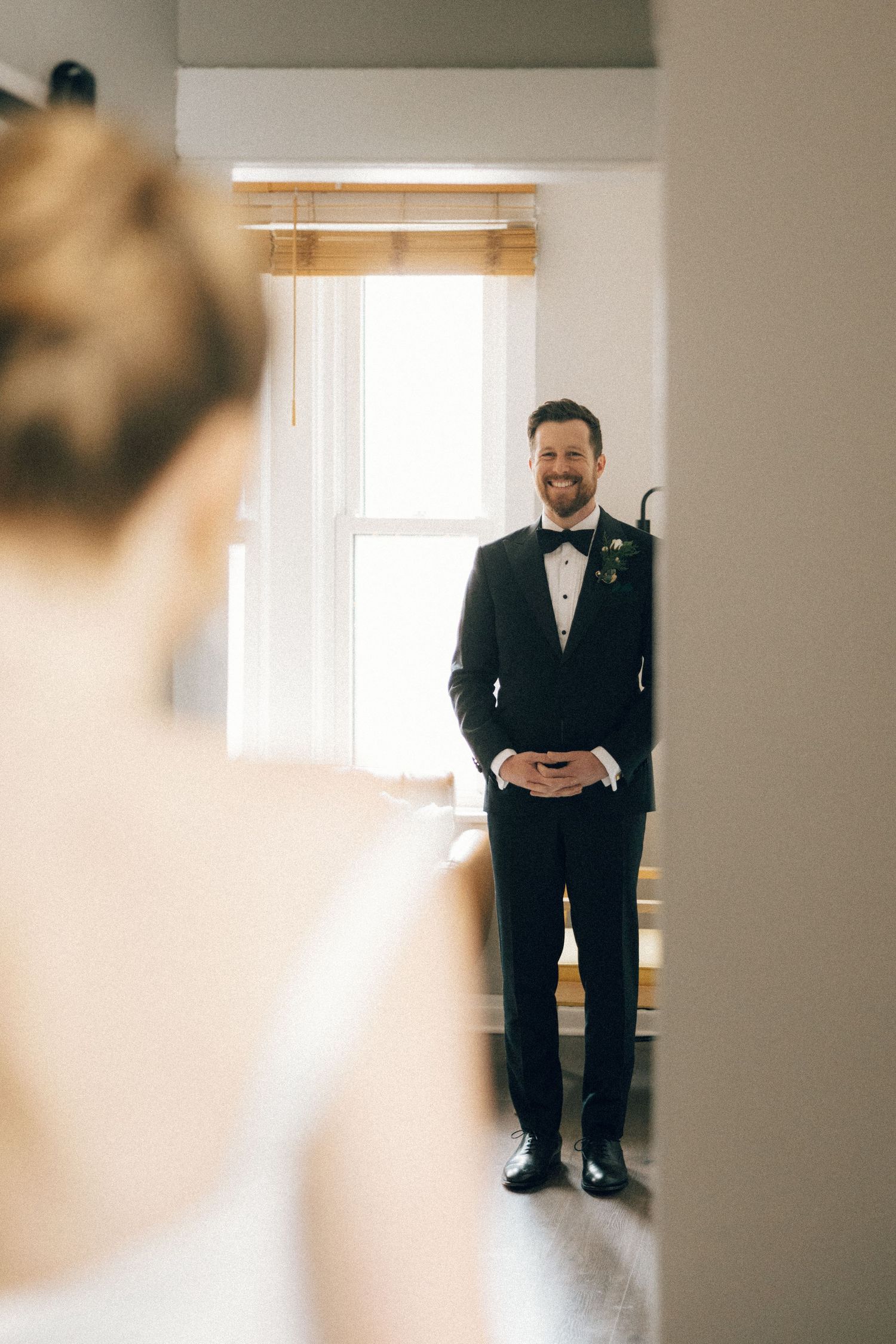 A groom in a classic black tuxedo stands in a bright room with natural window light.