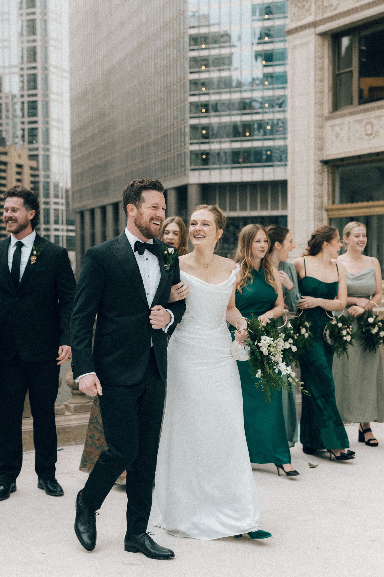 Wedding party in formal attire poses outside modern glass building with bridesmaids in emerald green dresses.