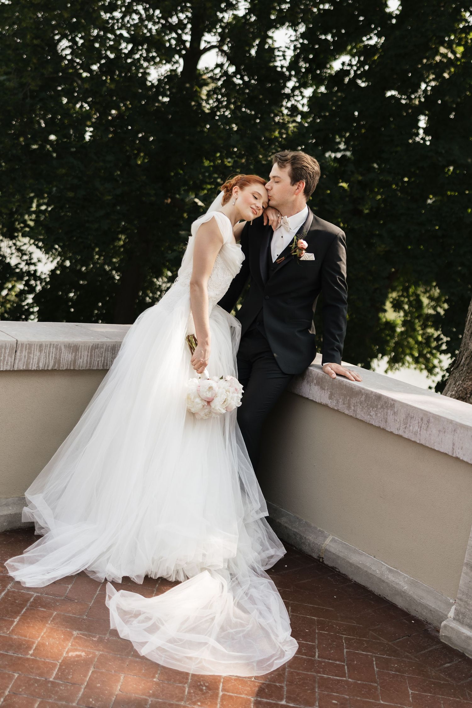 Bride and groom share a romantic kiss on a balcony, bride in white tulle gown holding bouquet, groom in black suit.