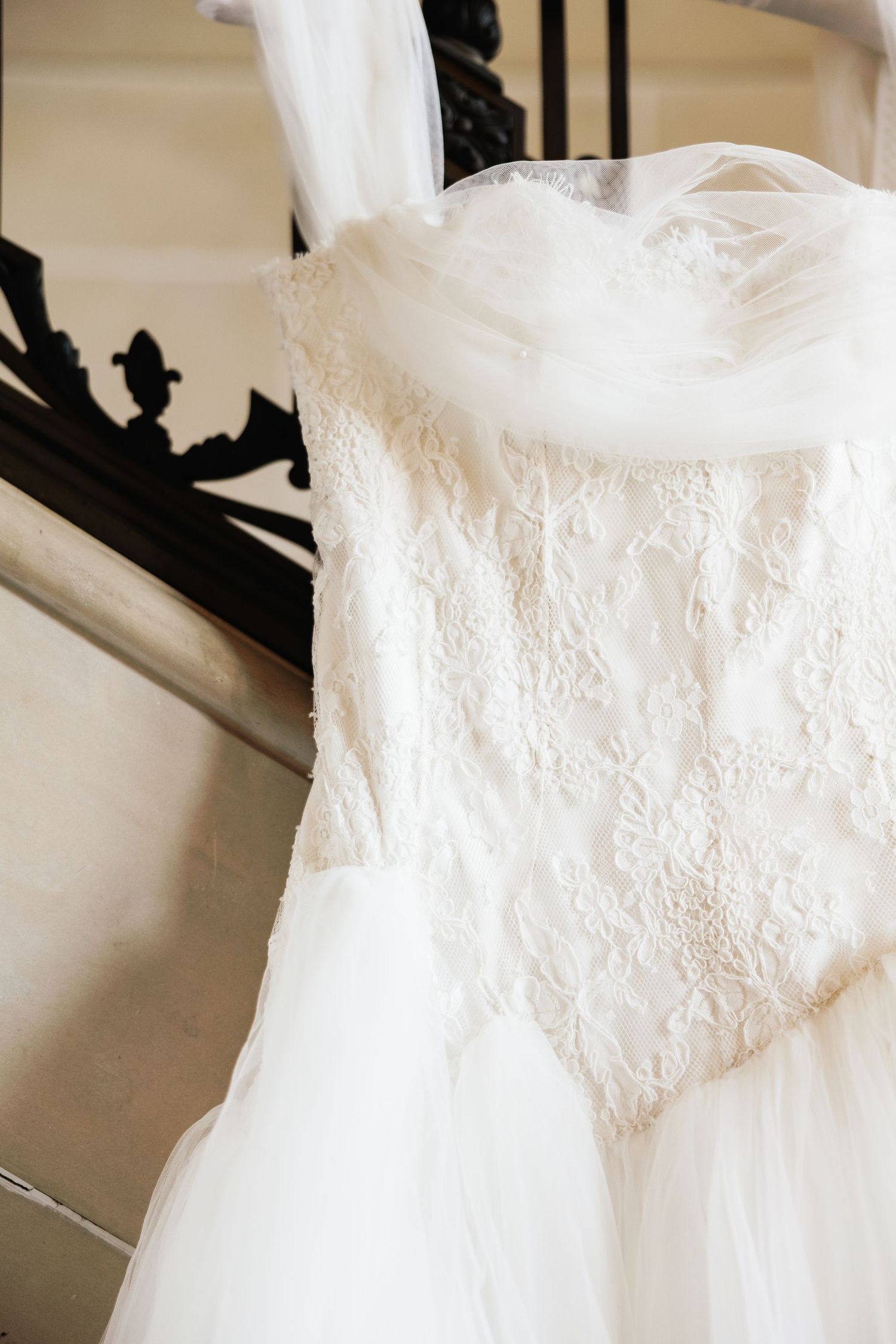 White lace wedding dress with tulle veil hanging on ornate black staircase railing in elegant venue.