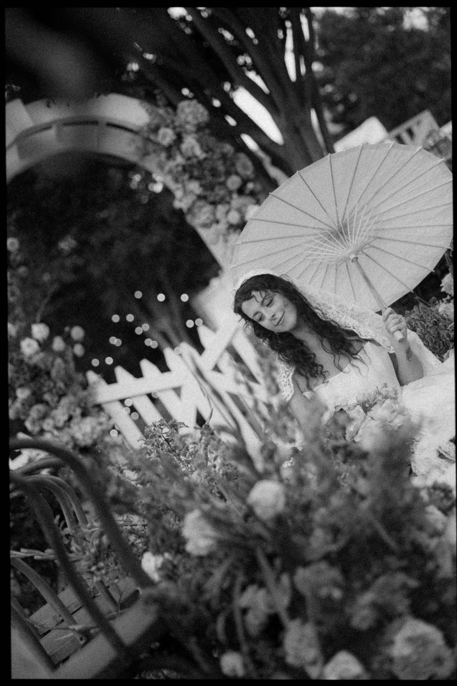 Black and white photo of a woman with long hair holding a parasol umbrella among lush floral arrangements outdoors.