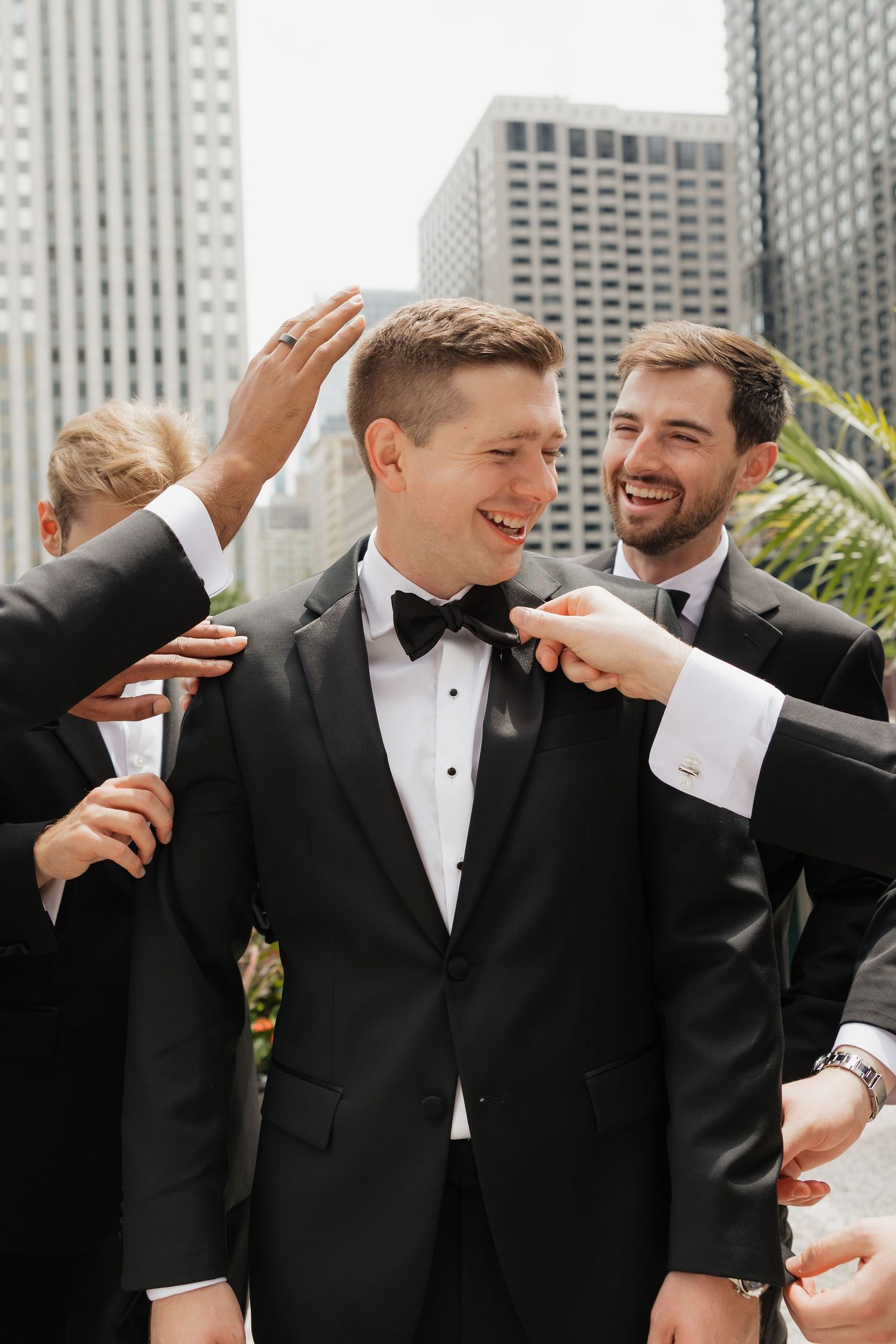 Groomsmen in black tuxedos help adjust the groom's bow tie on a rooftop with city skyscrapers in the background.