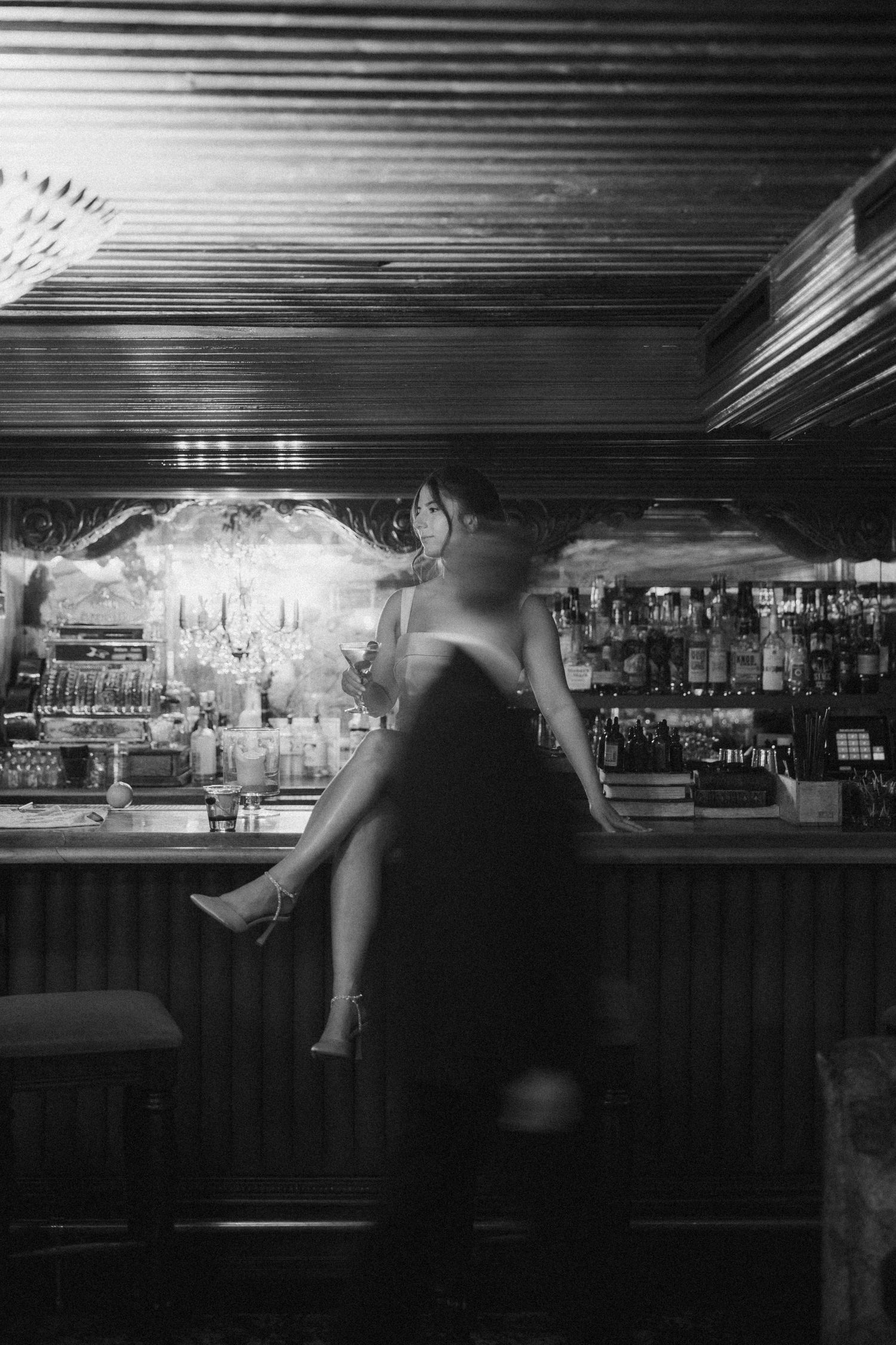Artistic black and white photo of a figure in a little black dress sitting at an illuminated bar counter.
