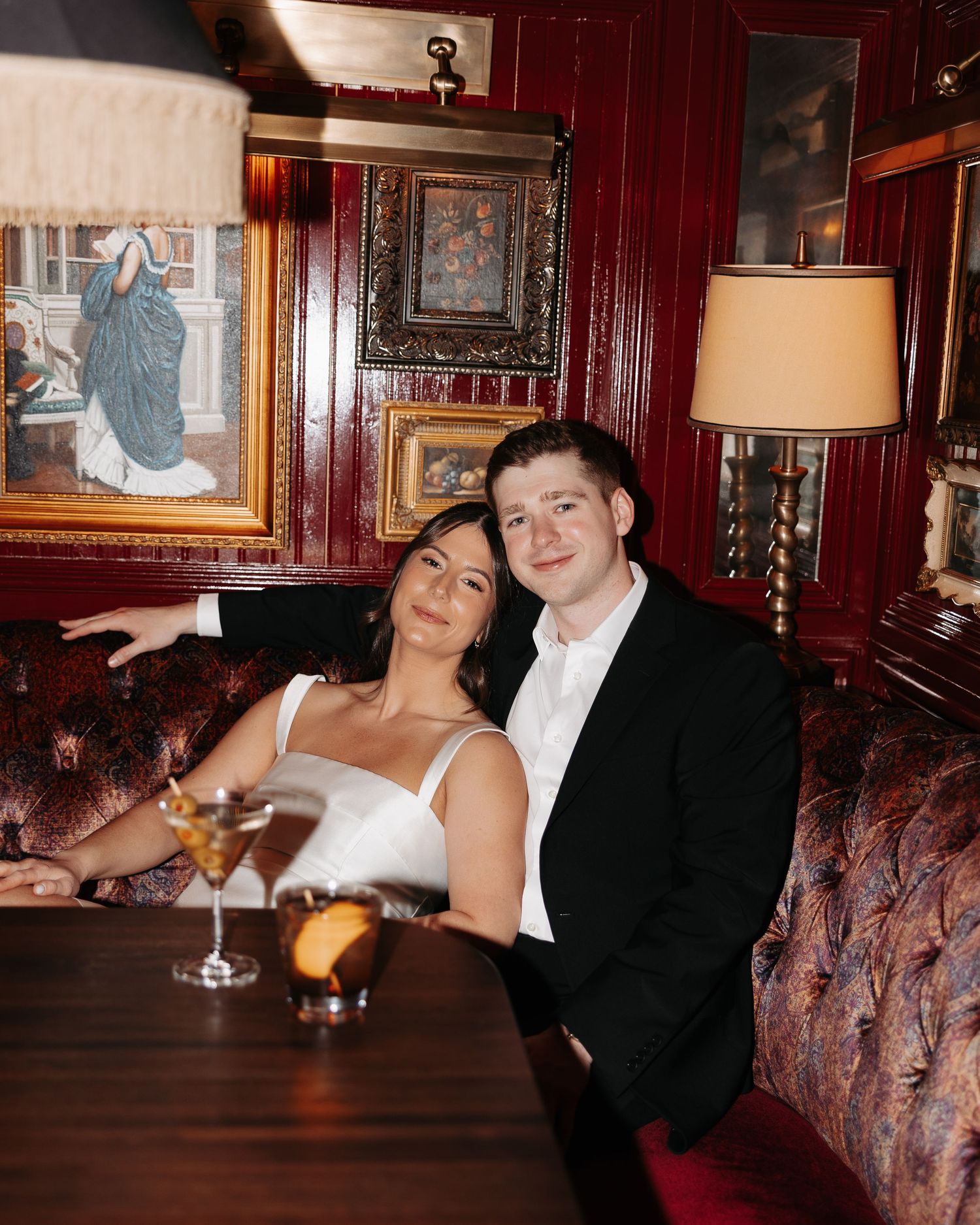 A couple in formal attire relaxes on a red leather couch in an elegant bar setting with wood paneling.