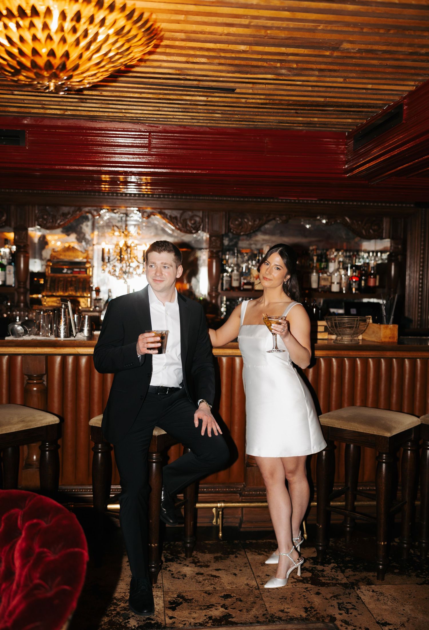Stylish pair sharing cocktails while standing at a retro bar counter with ornate ceiling fixture above.