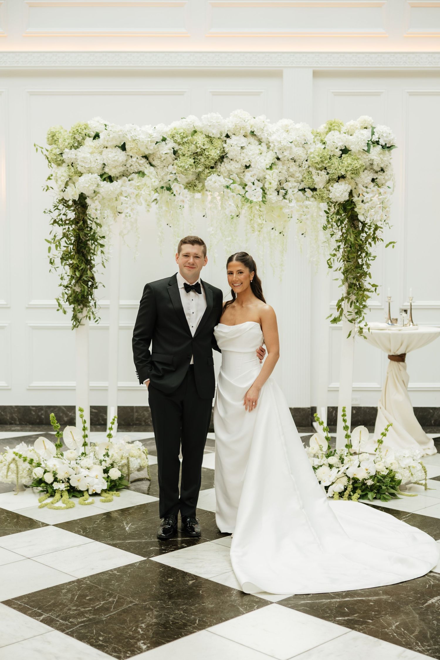 Bride and groom pose under a white floral arch with cascading greenery on a checkered marble floor.