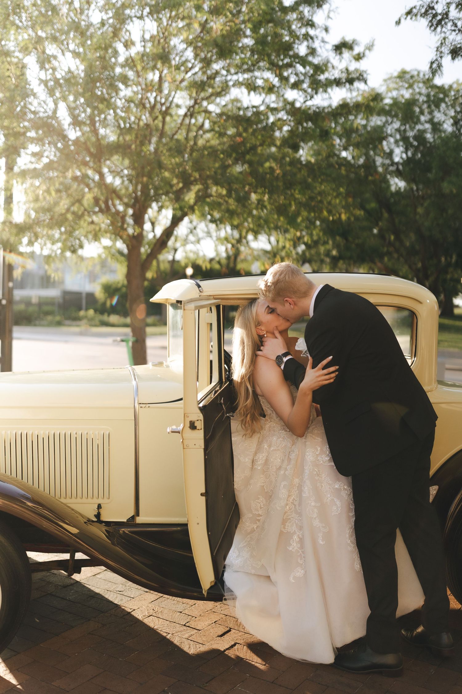 Romantic moments captured beside a vintage cream-colored car under leafy trees.