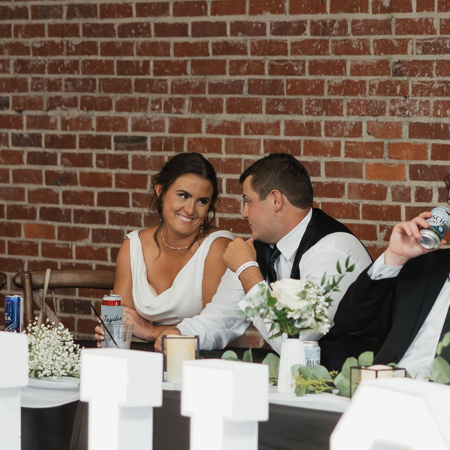 Wedding reception table setting with brick wall backdrop shows guests seated at white-clothed tables.