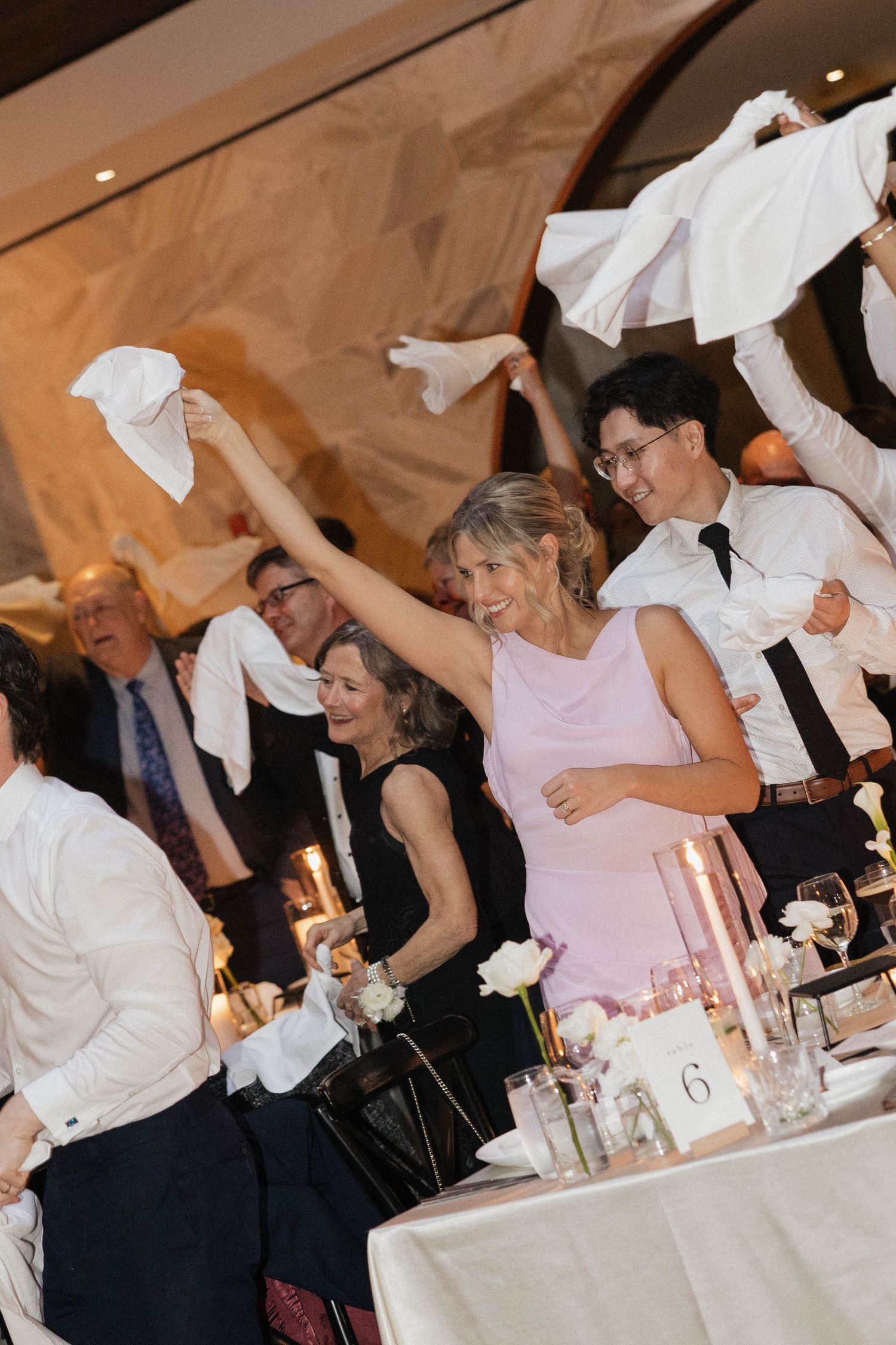 Guests wave white napkins in celebration at an elegant wedding reception dinner, with candles and floral centerpieces on table 6.