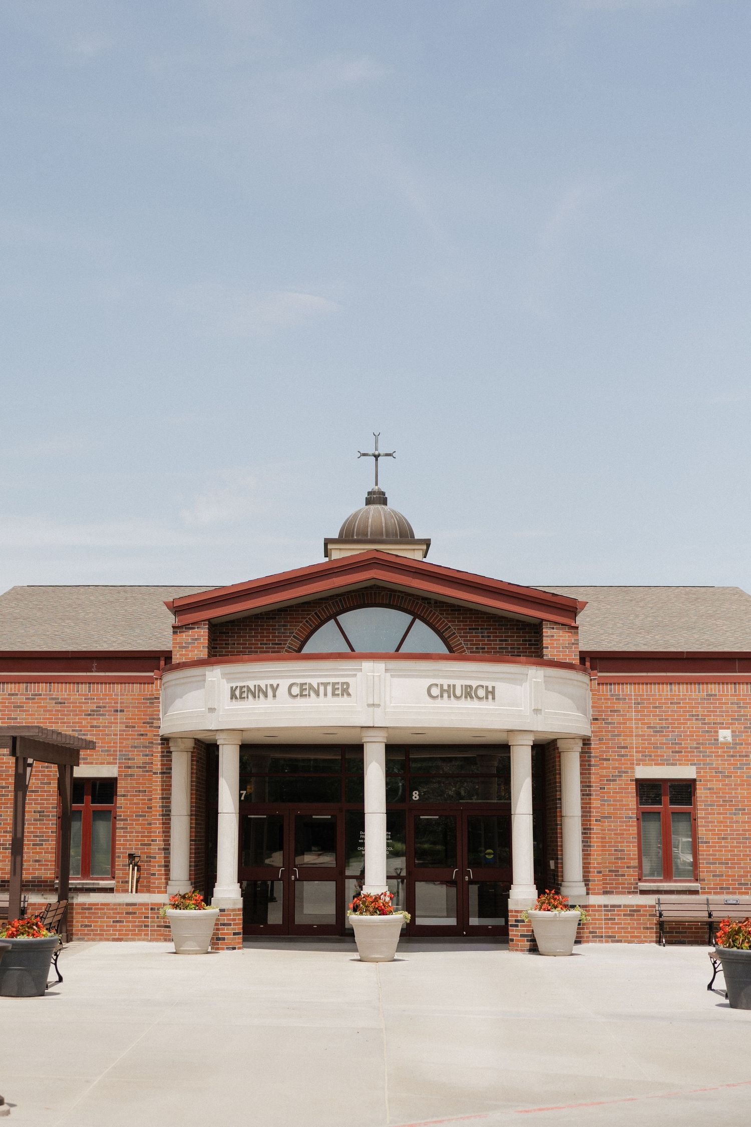 Brick exterior of Kenny Center Church with white columns, arched entrance, cross on dome, and flower planters on a sunny day.