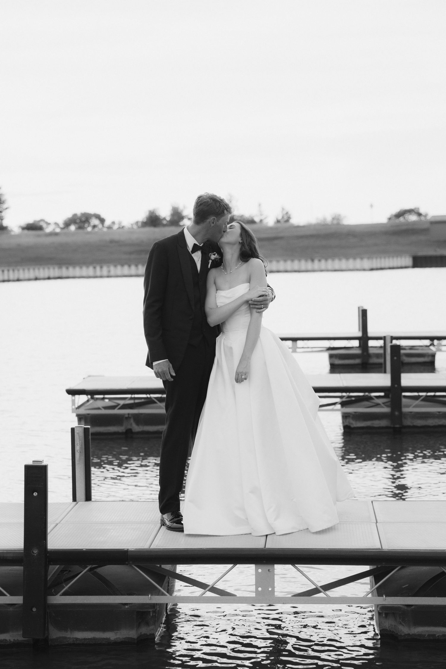 Black and white photo of a bride and groom sharing a romantic kiss on a wooden dock by the water.
