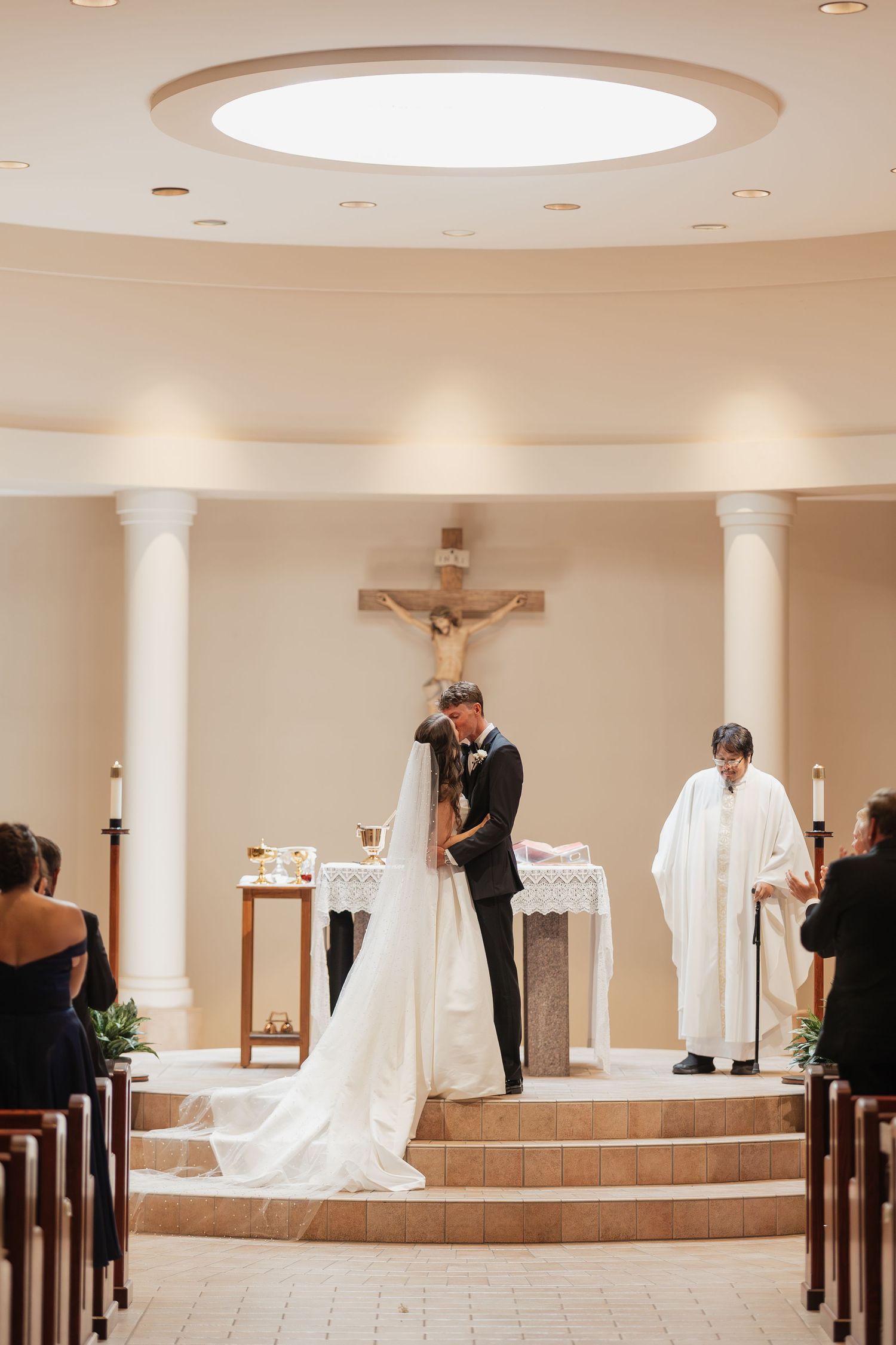 Bride and groom share first kiss at Catholic church altar with priest and crucifix during elegant wedding ceremony.
