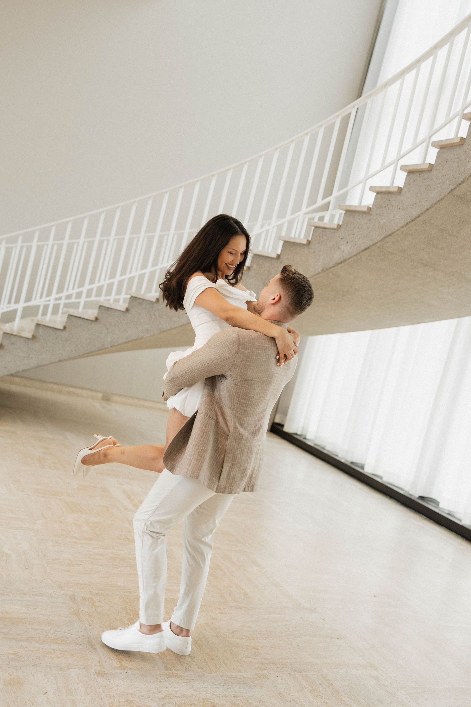 A man lifts a smiling woman in a bright, modern space with a curved staircase in the background.