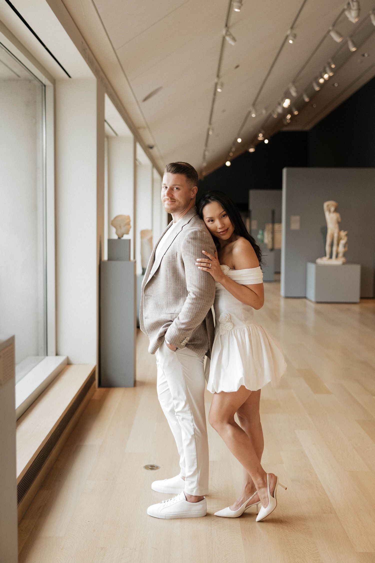 Couple posing in art museum gallery, woman in white mini dress embracing man in grey suit near ancient sculptures.