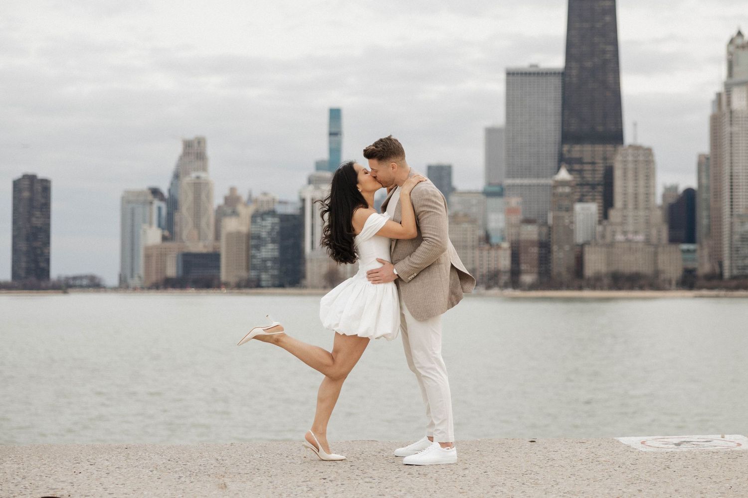 Couple kissing on Chicago waterfront, woman lifting leg, city skyline behind them.