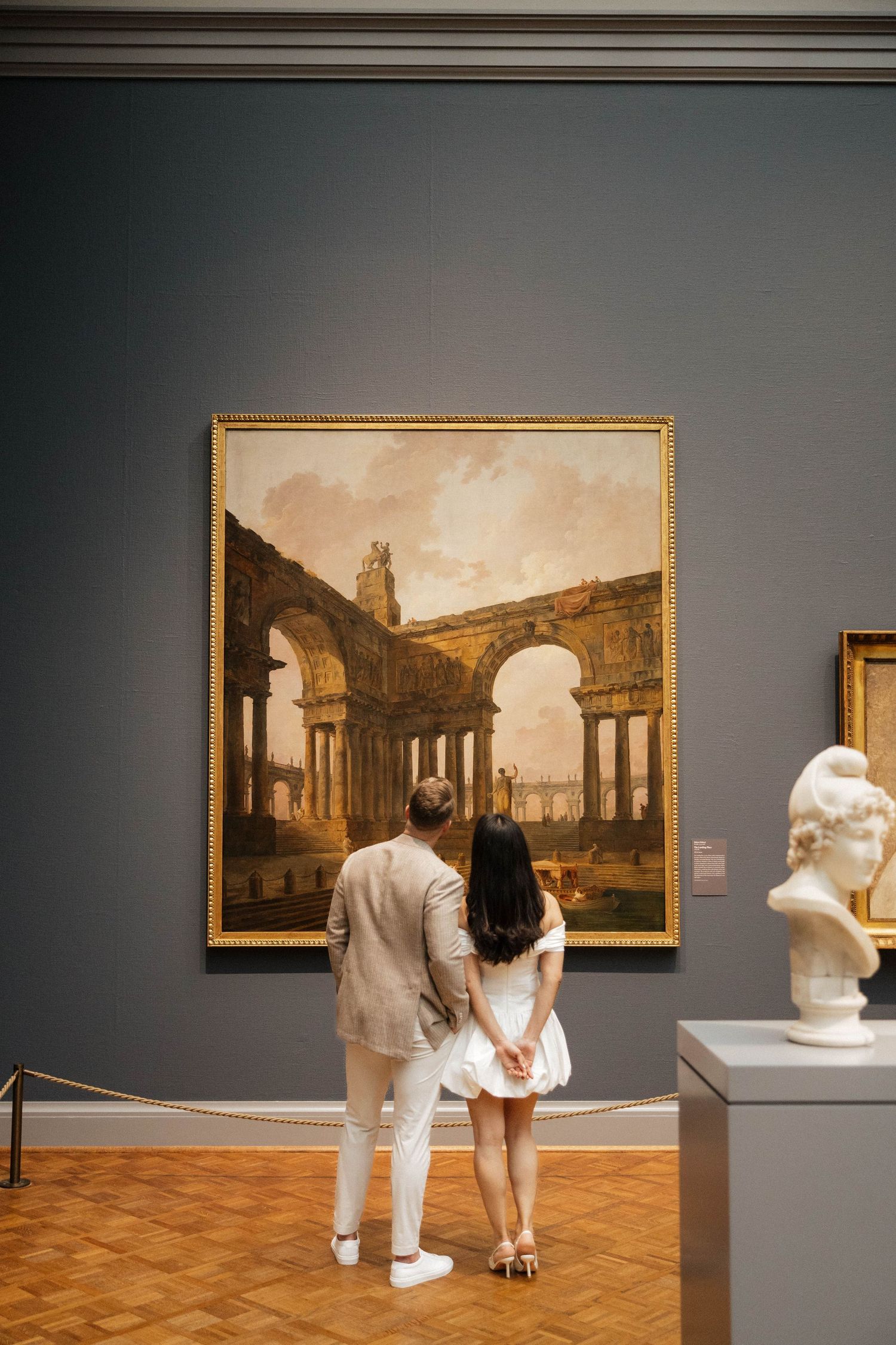 Couple viewing a classical architectural ruins painting in an art museum gallery with a marble bust.