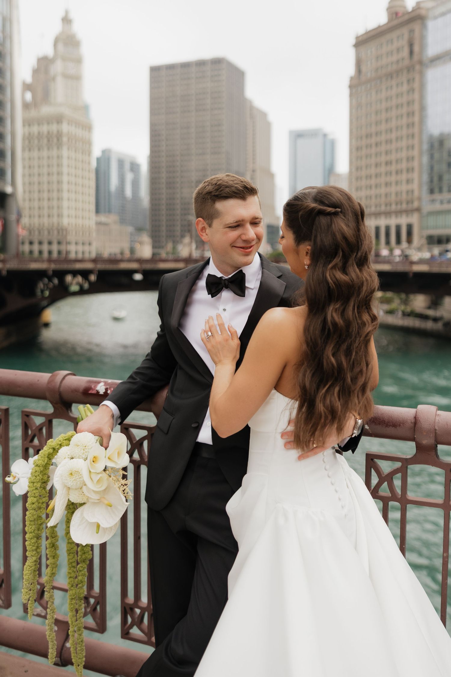 Smiling groom in a black tuxedo and bow tie embraces his bride in a white gown beside a city river, with a calla lily bouquet nearby.