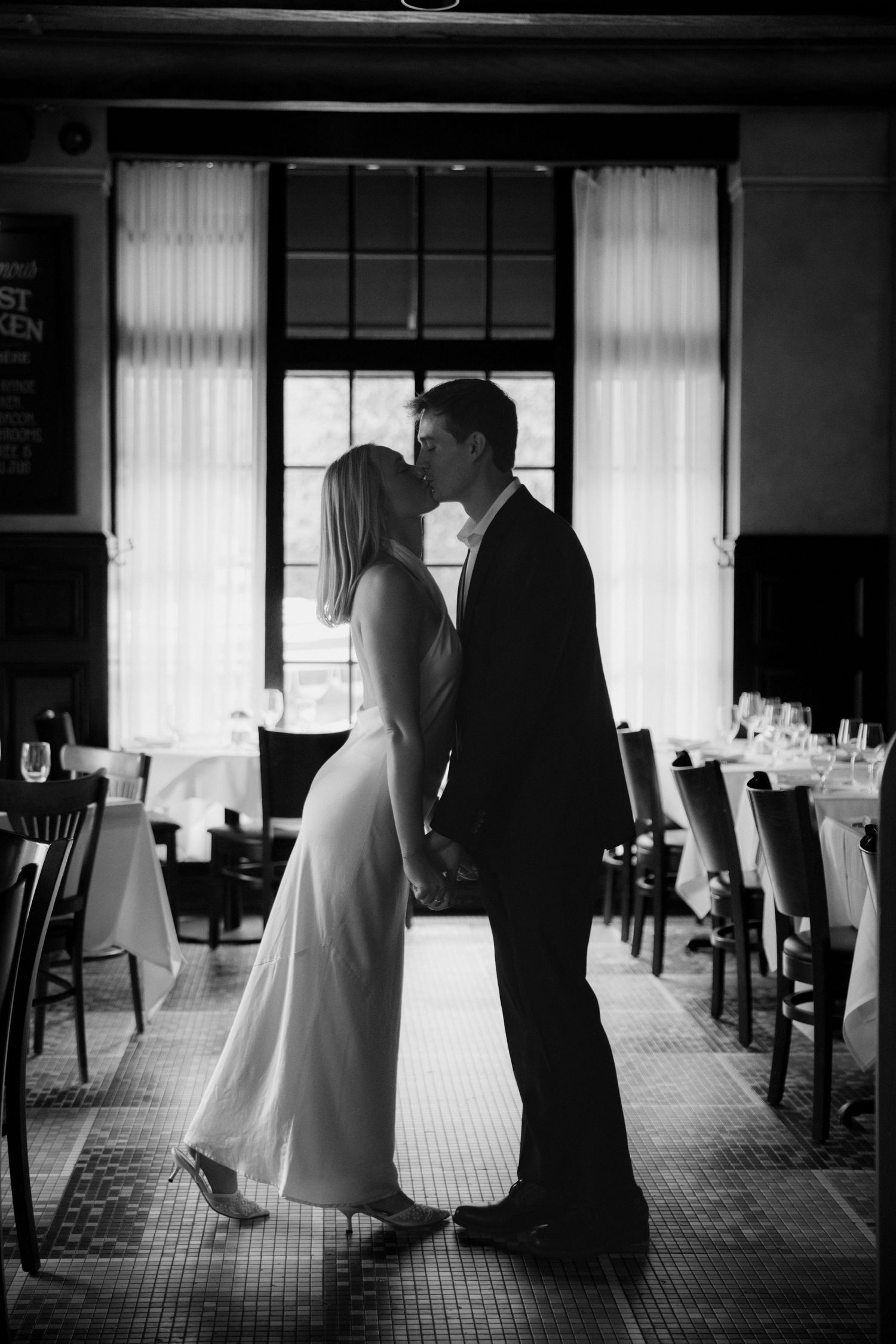Black and white photo of a couple kissing in an elegant restaurant, the woman in a long white dress and the man in a dark suit.
