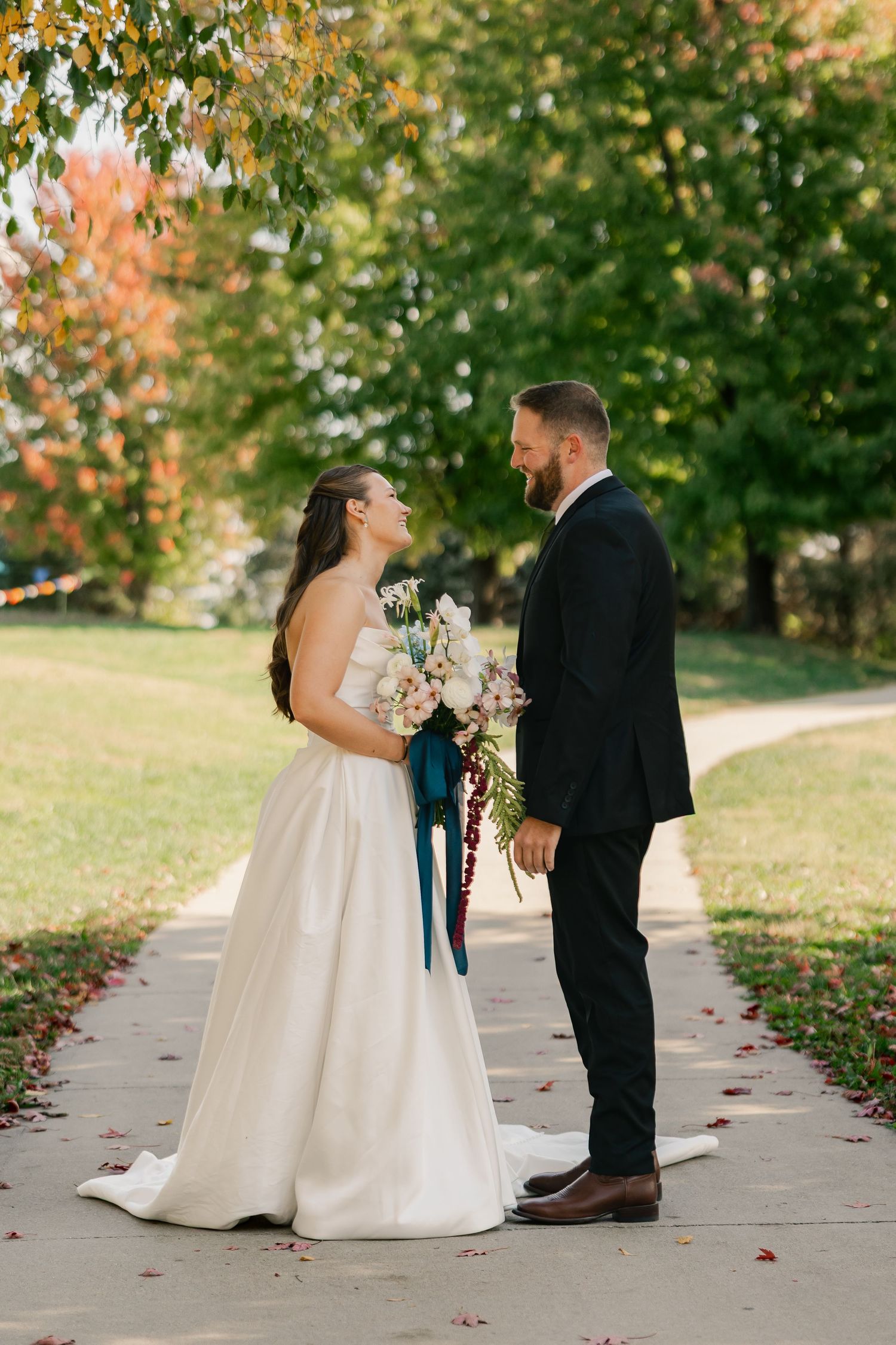 Bride in a white satin gown holding a teal-ribboned bouquet faces her bearded groom in a dark suit on a leaf-covered path in autumn.