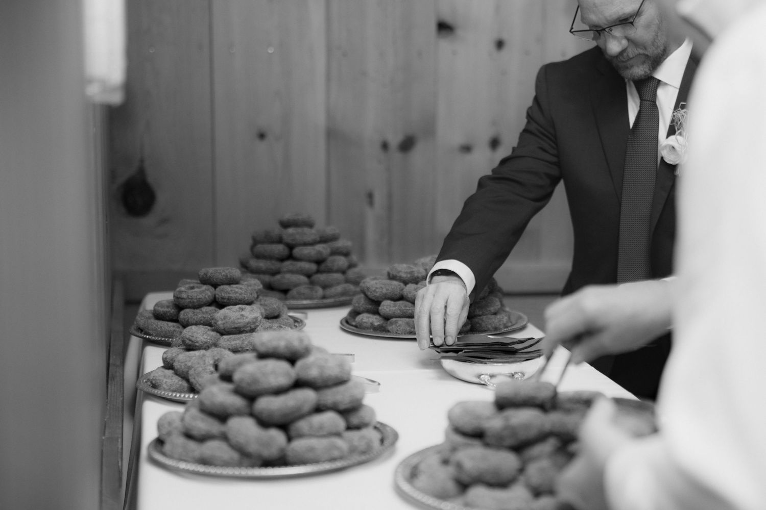 Black and white photo of a man in a suit and glasses serving golden croquettes from stacked platters on a buffet table at an event.