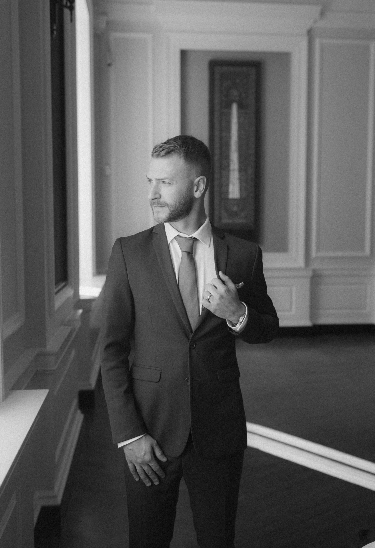 Black and white artistic wedding photo showing groom in suit looking contemplatively out a bright window.