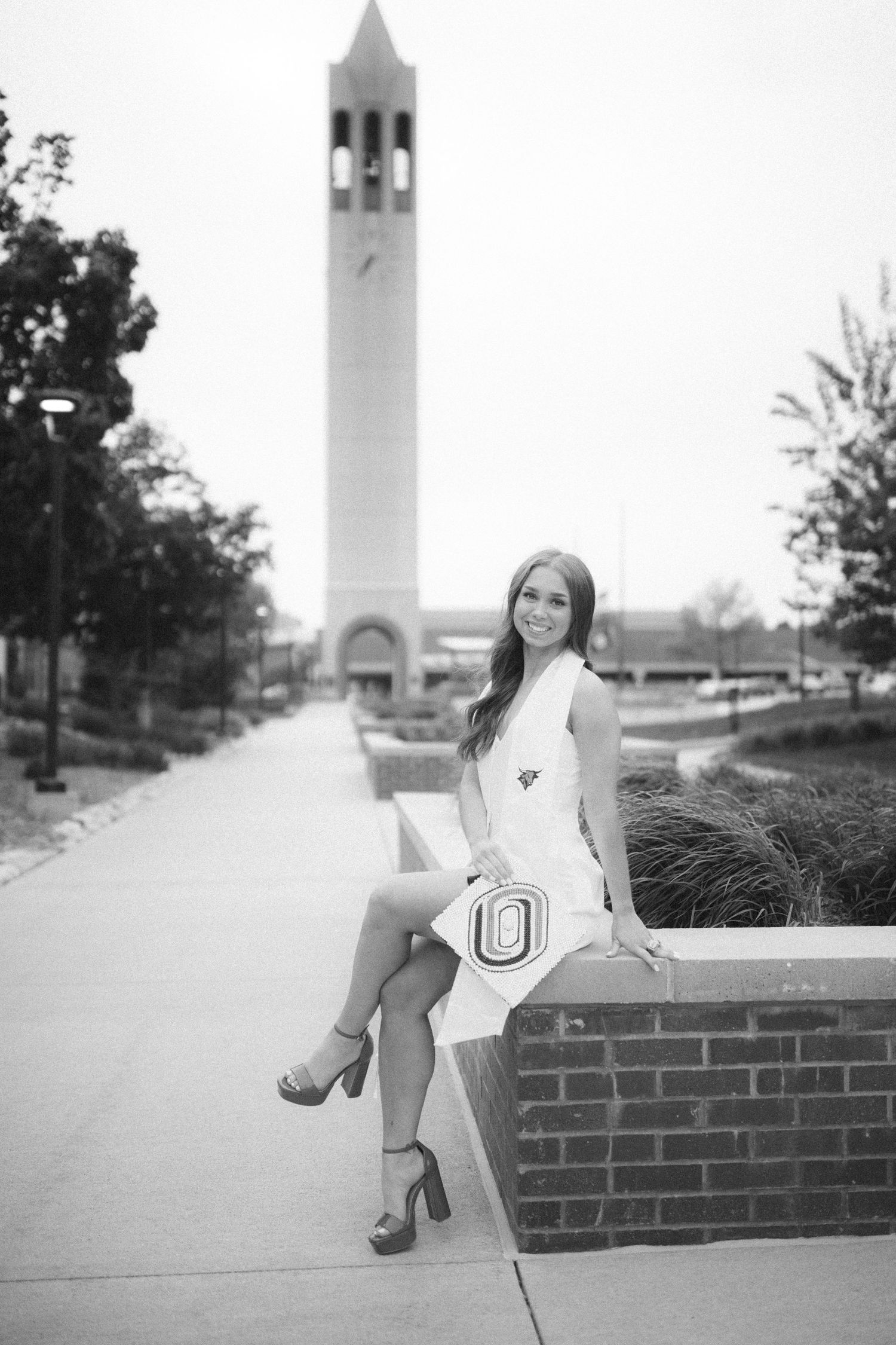Black and white photo of someone in a white dress sitting on a brick wall with a tall bell tower in the background.