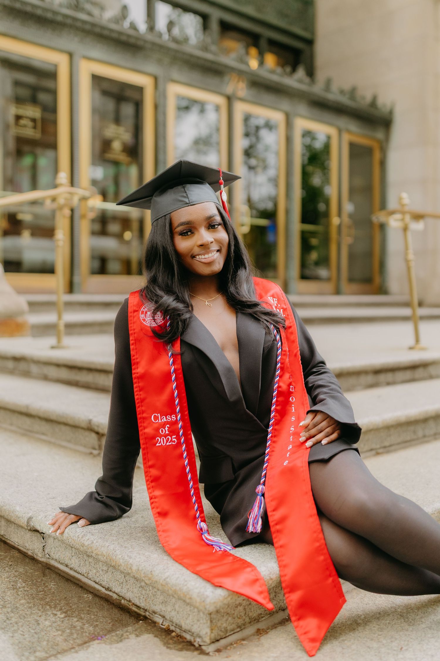 Graduate in cap and gown with red graduation stole poses on stone steps during outdoor photo session.