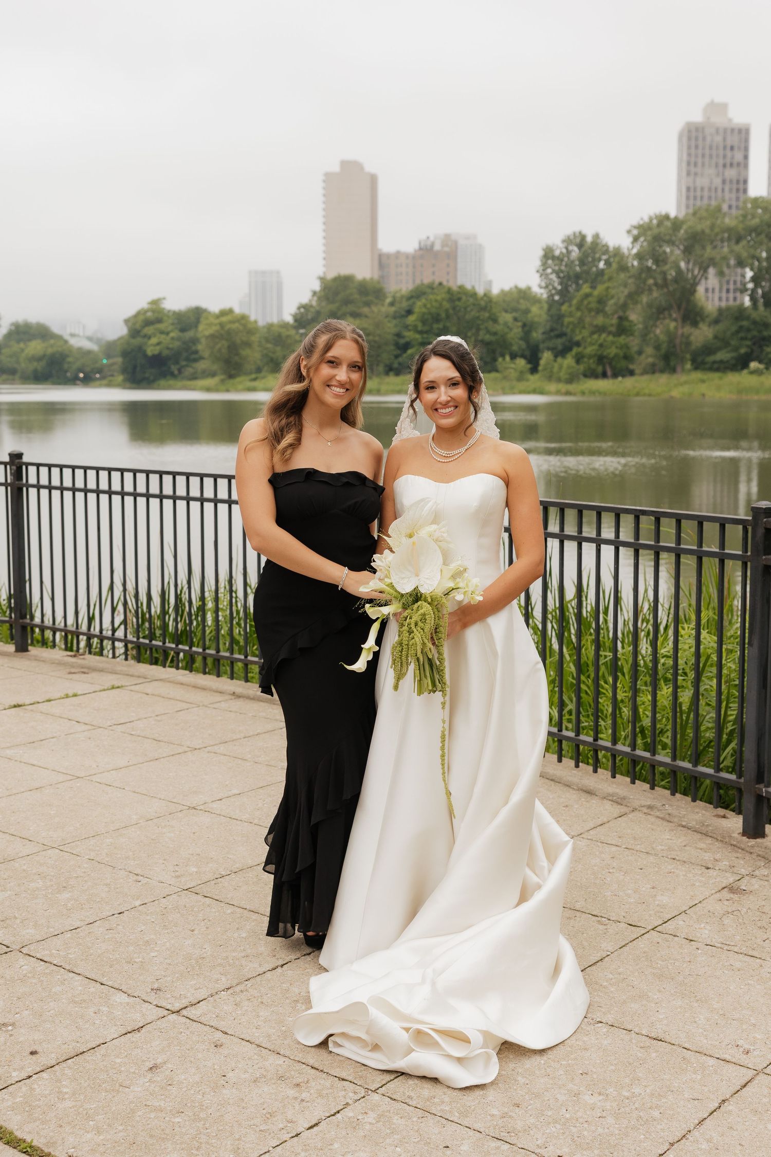 Bride in white gown holding calla lily bouquet poses with bridesmaid in black dress by a lake in a city park.