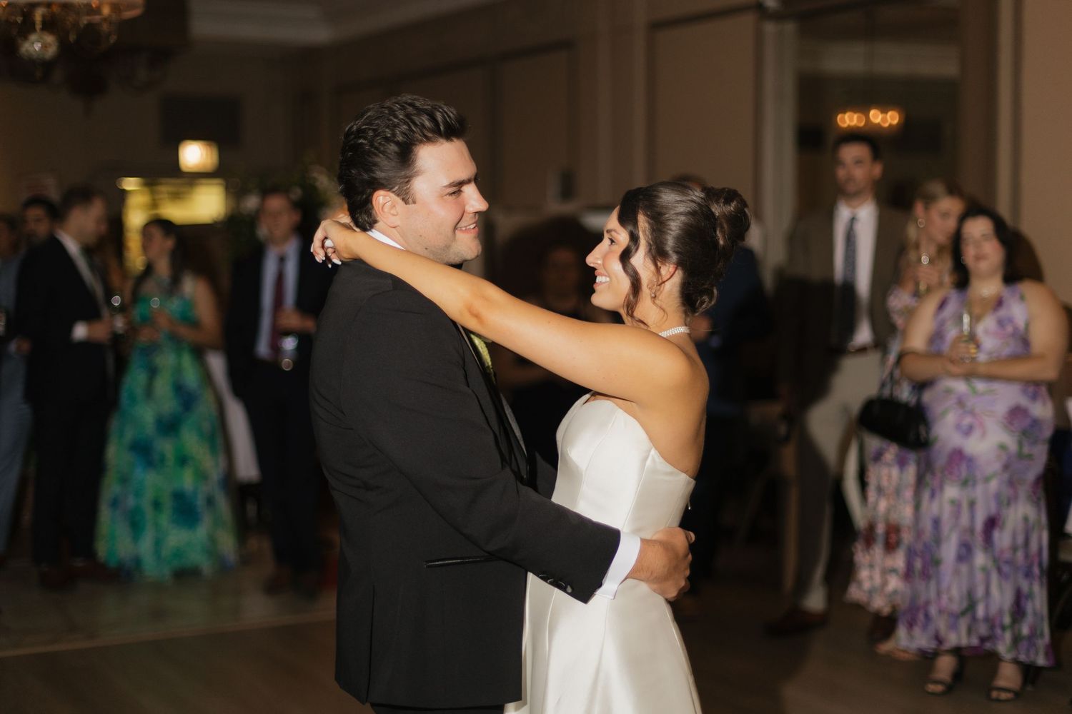 Bride and groom share their first dance at elegant wedding reception with guests watching in background.