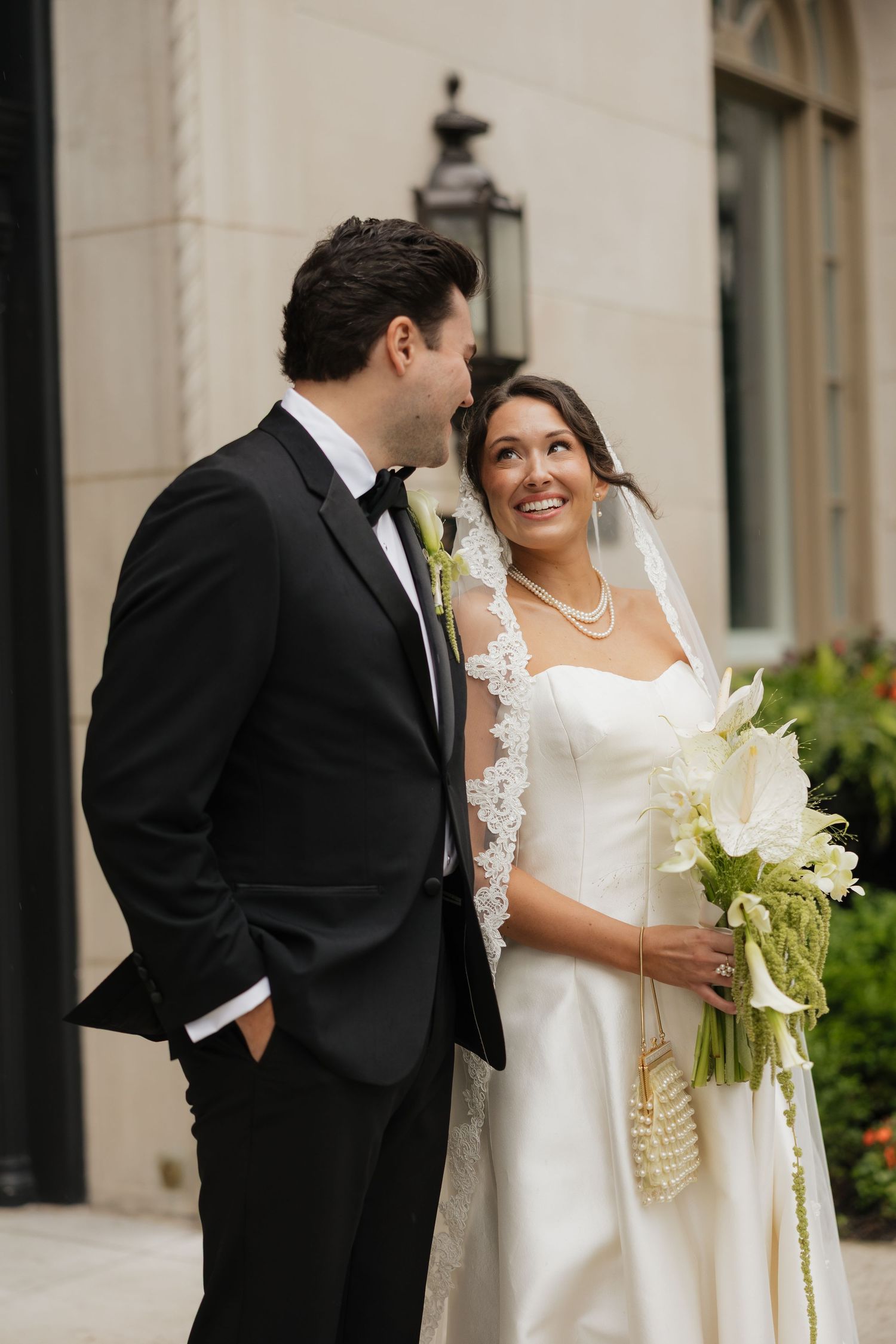 Bride in white gown holds calla lily bouquet, smiling at groom in black tuxedo outside elegant building.