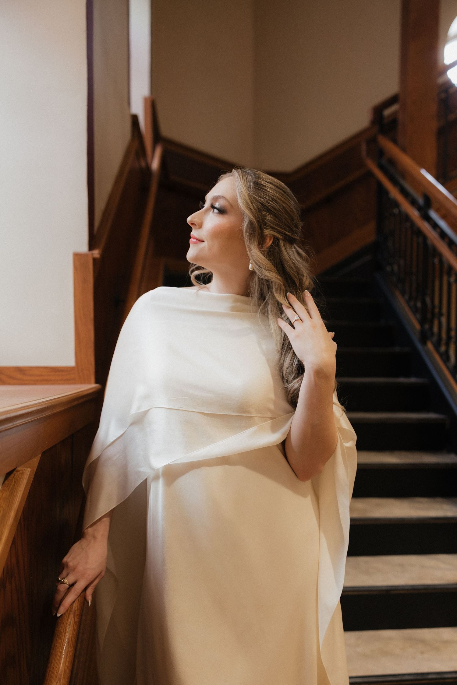 Elegant white wedding dress with flowing cape sleeves photographed on a staircase in soft natural light.